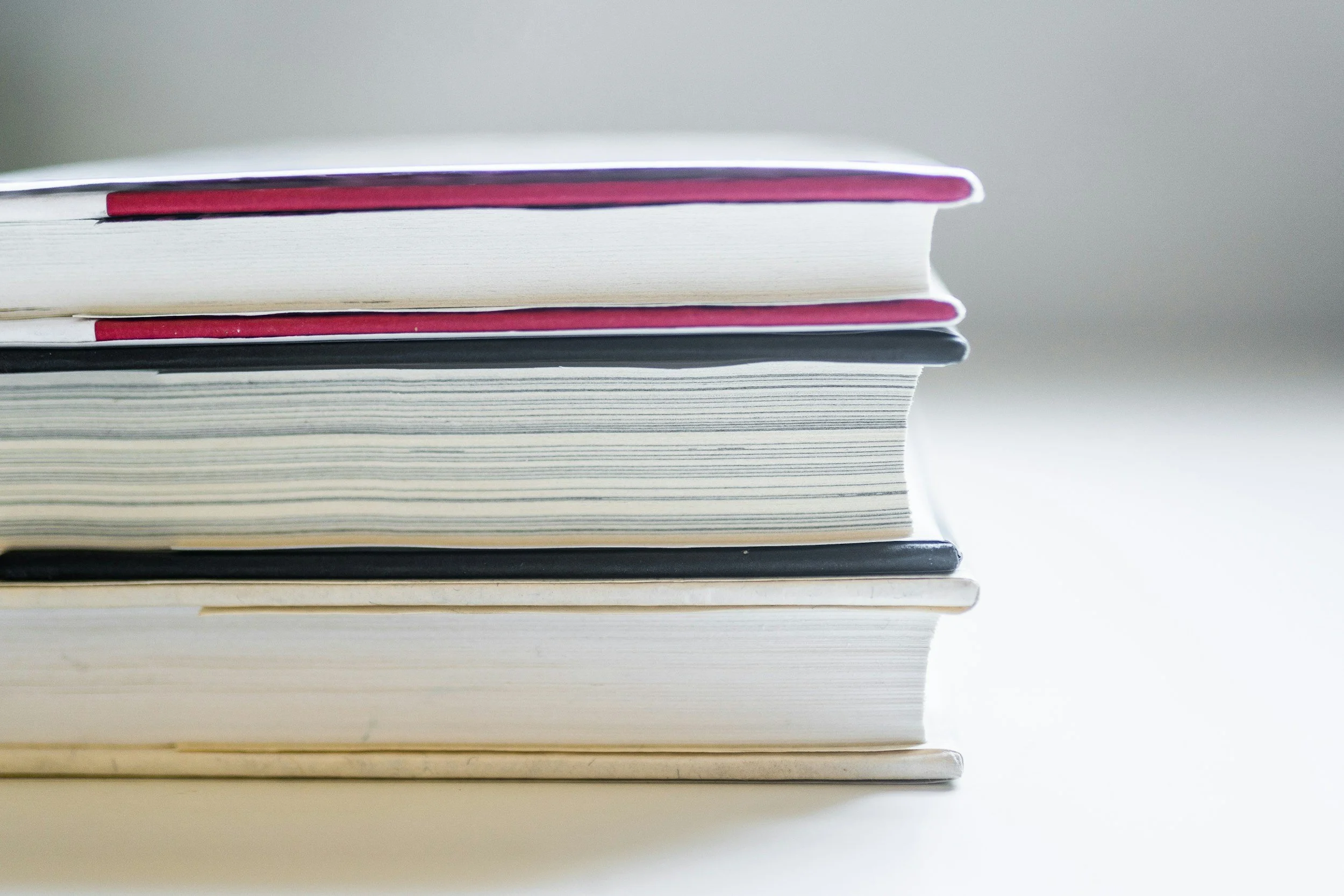 Close-up of a stack of books or notebooks with various colored covers, arranged horizontally on a white surface.