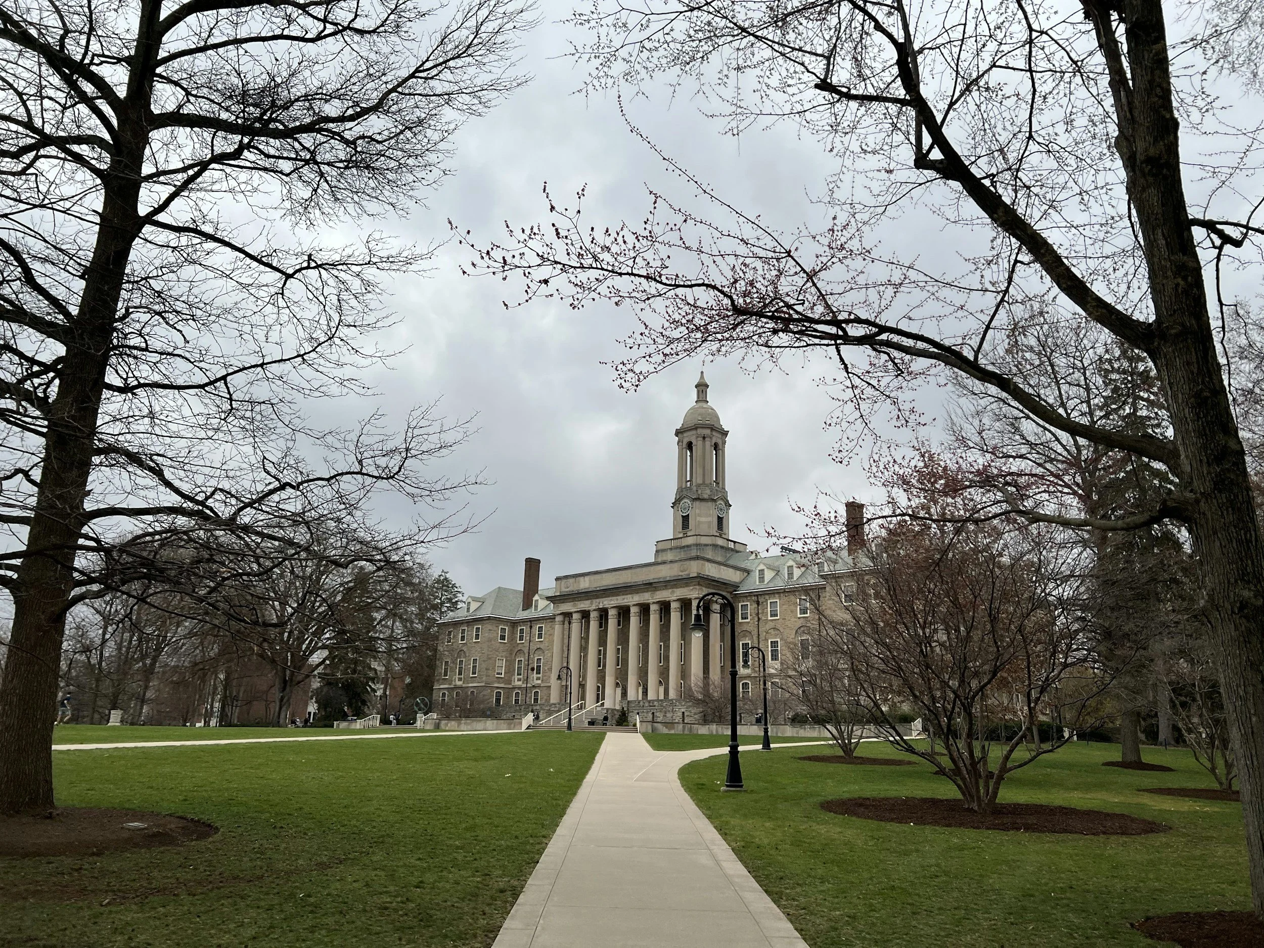View of a college campus or government building with a prominent bell tower, surrounded by trees with no leaves and a well-maintained lawn, overcast sky.