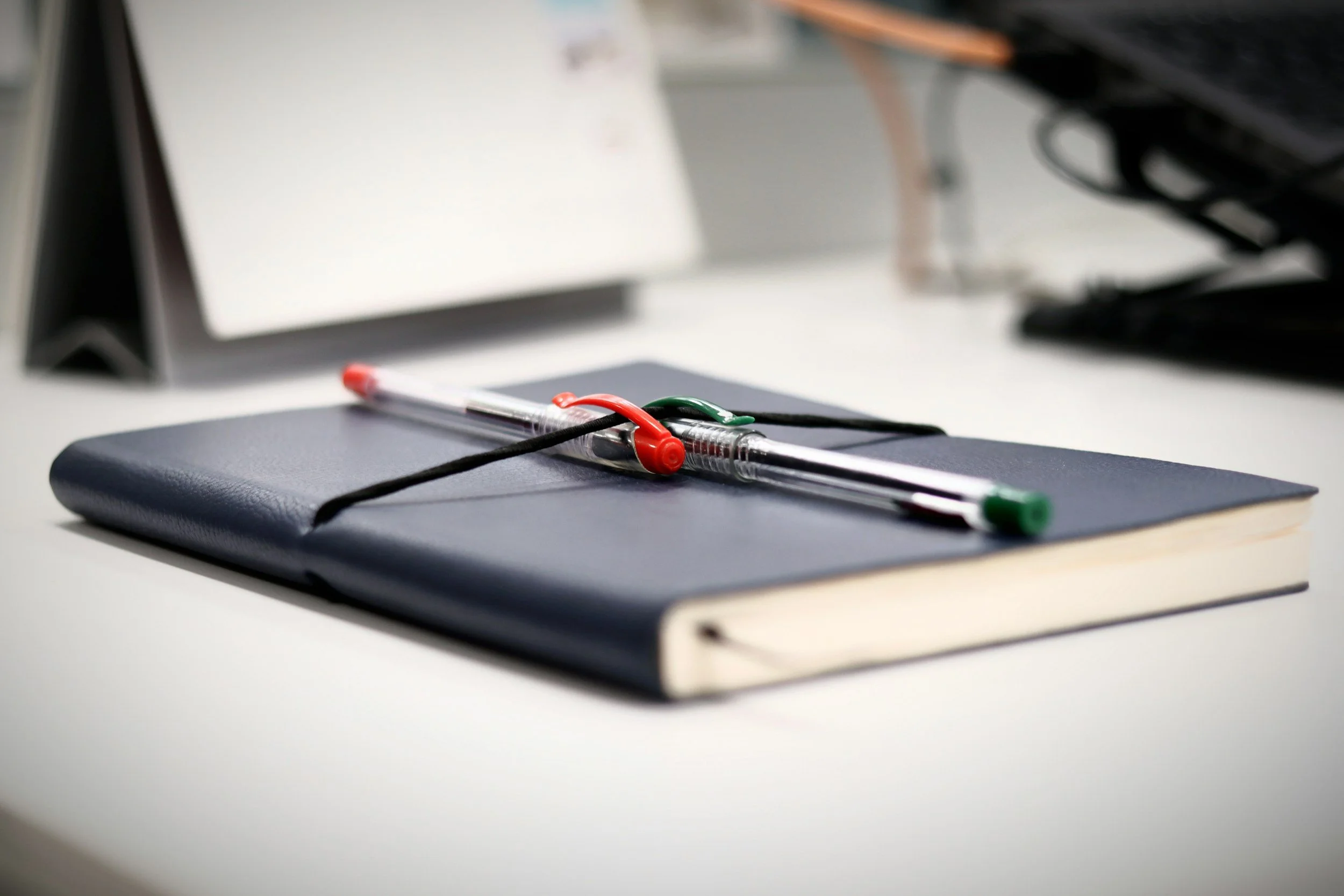 Close-up of a black notebook with a black elastic strap, on top of which a clear plastic pen with a red and green cap is placed, on a white surface. Blurred background includes a white panel and some office supplies.