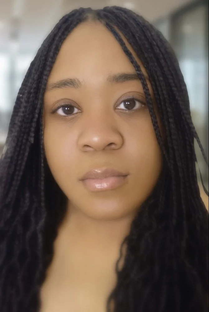 Close-up of a young woman with long, dark braids, looking directly at the camera with a neutral expression, indoors with a blurred background.