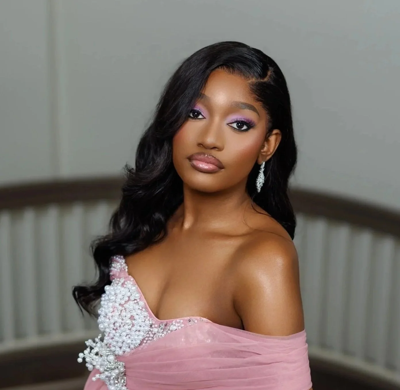 A woman with dark hair pulled back, wearing large gold earrings and a white sleeveless top, posed with her hand near her face, in front of a light-colored background with green leaves.