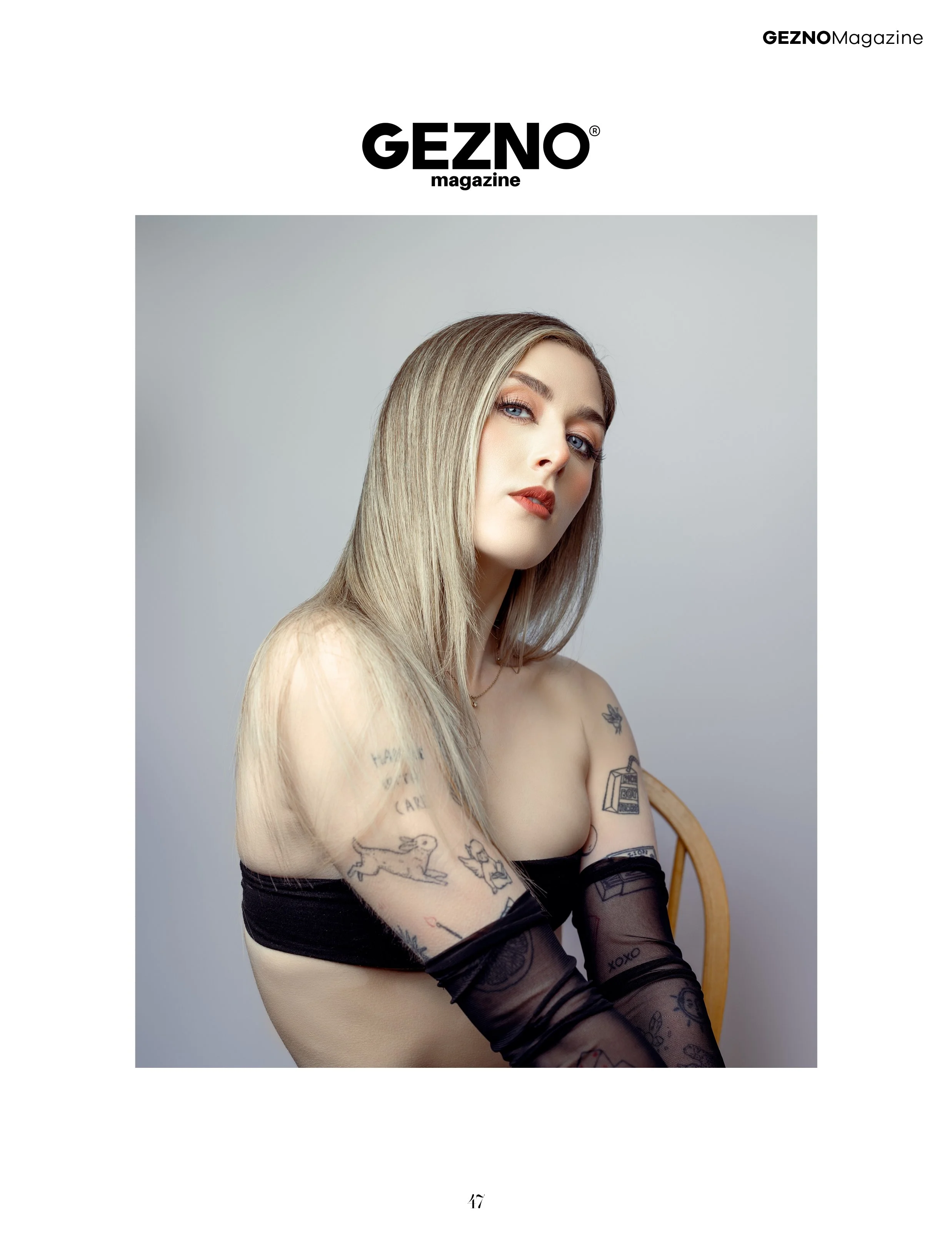 A woman with long blonde hair and tattoos, wearing a black strapless top and sheer black gloves, seated on a wooden chair against a plain light background, posing for a photo on the cover of GEZNO magazine.
