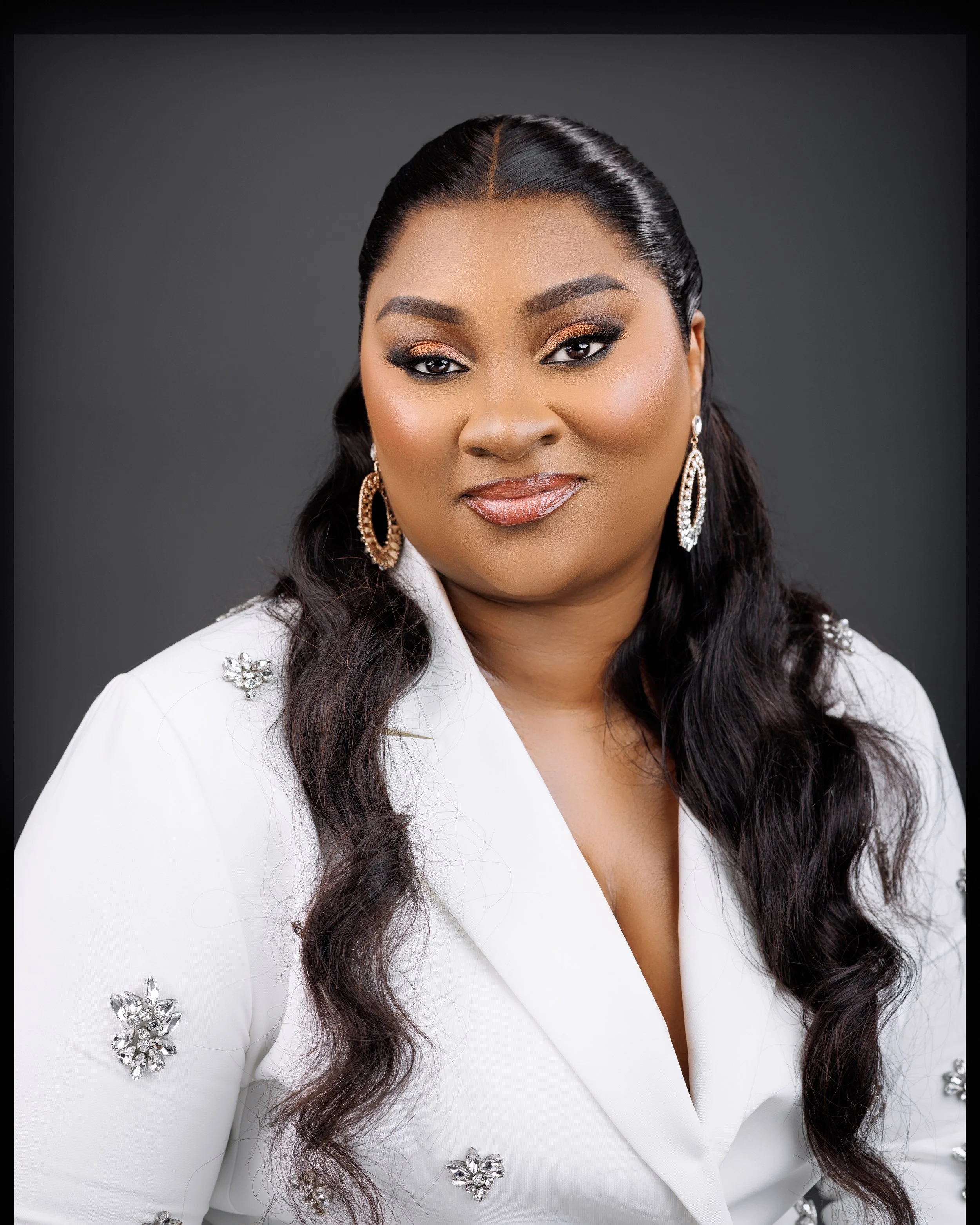 A woman with long wavy dark hair, wearing a white blazer with decorative embellishments and large, gold and silver earrings, smiling softly against a dark gray background.