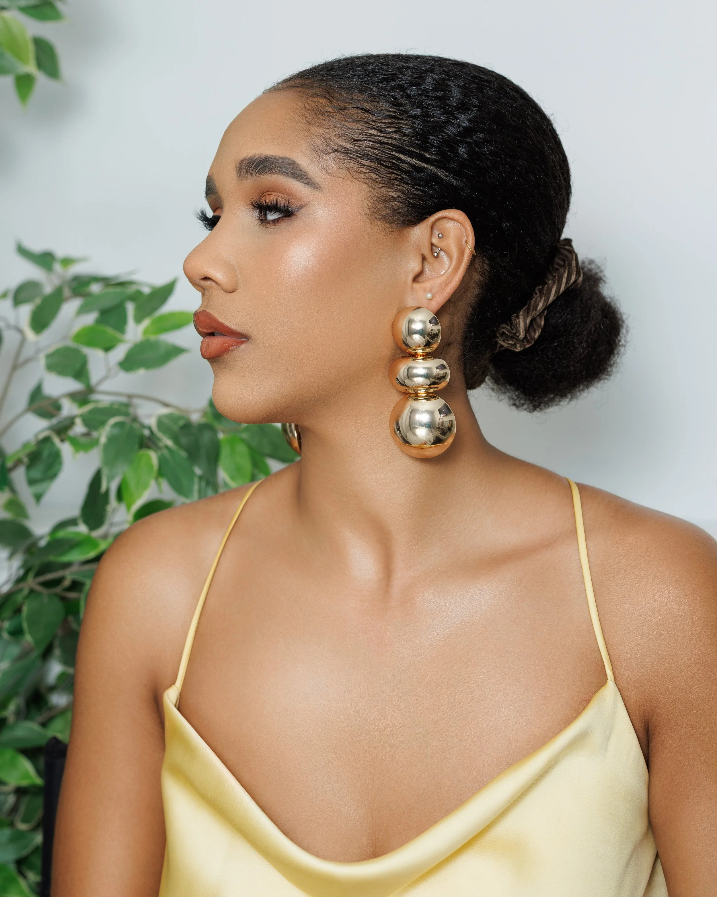Side profile of a woman with dark hair styled in a bun, large gold spherical earrings, wearing a yellow satin spaghetti strap top, standing next to a green leafy plant.