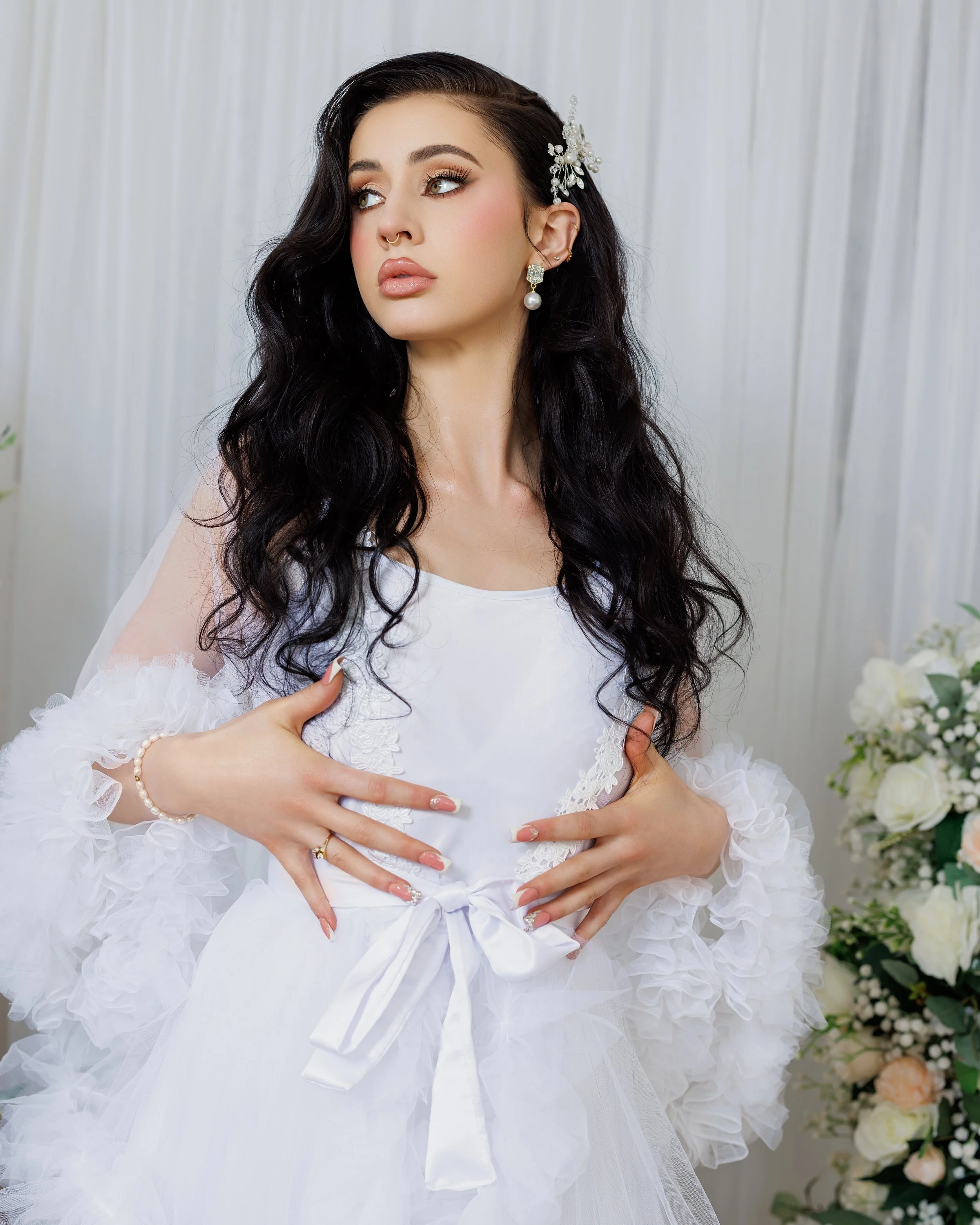 A woman with long dark hair wearing a white dress with lace and tulle, accessorized with pearl earrings, a pearl bracelet, a floral hairpiece, and makeup, standing in front of a white curtain with flowers on the side.