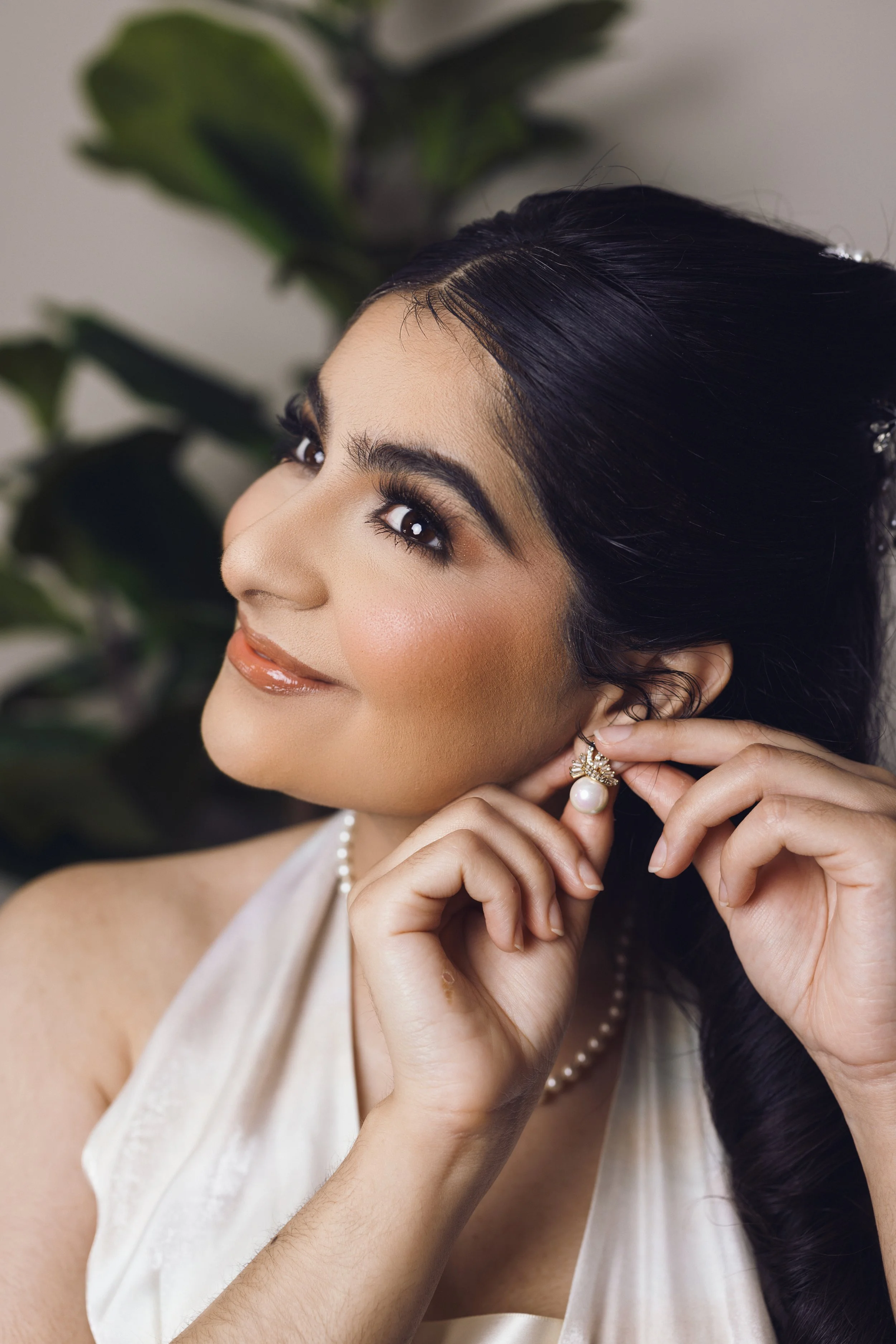 A woman with dark hair and makeup, wearing pearl earrings and a pearl necklace, is putting on an earring. She is smiling slightly and looking upward, with a blurred green plant in the background.