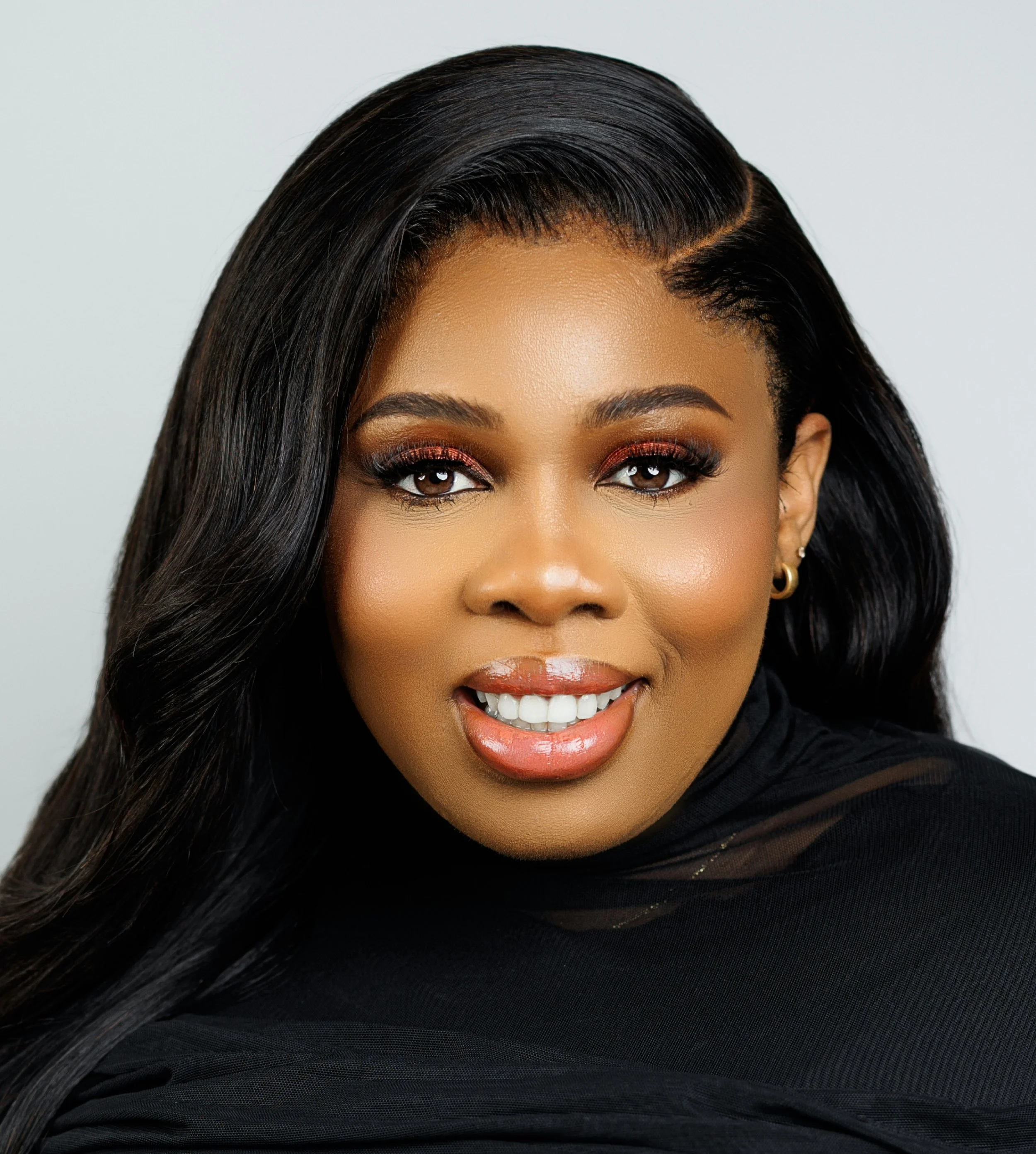 Close-up portrait of an African American woman with long black hair, makeup, gold earrings, and a black top, smiling at the camera.