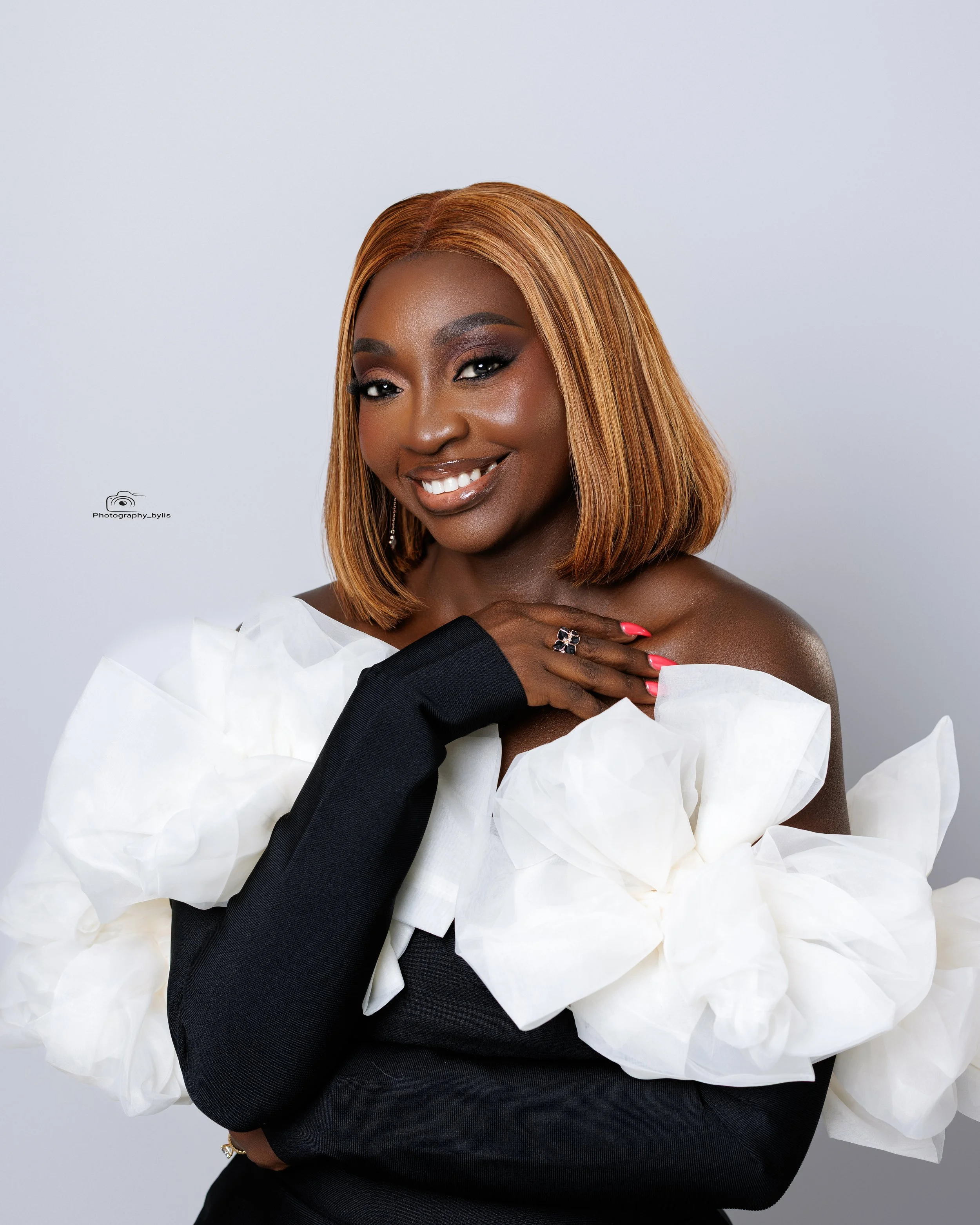 Portrait of a woman with shoulder-length blonde hair, wearing a black top with large white ruffled sleeves, smiling and posing with her hand on her chest against a plain white background.