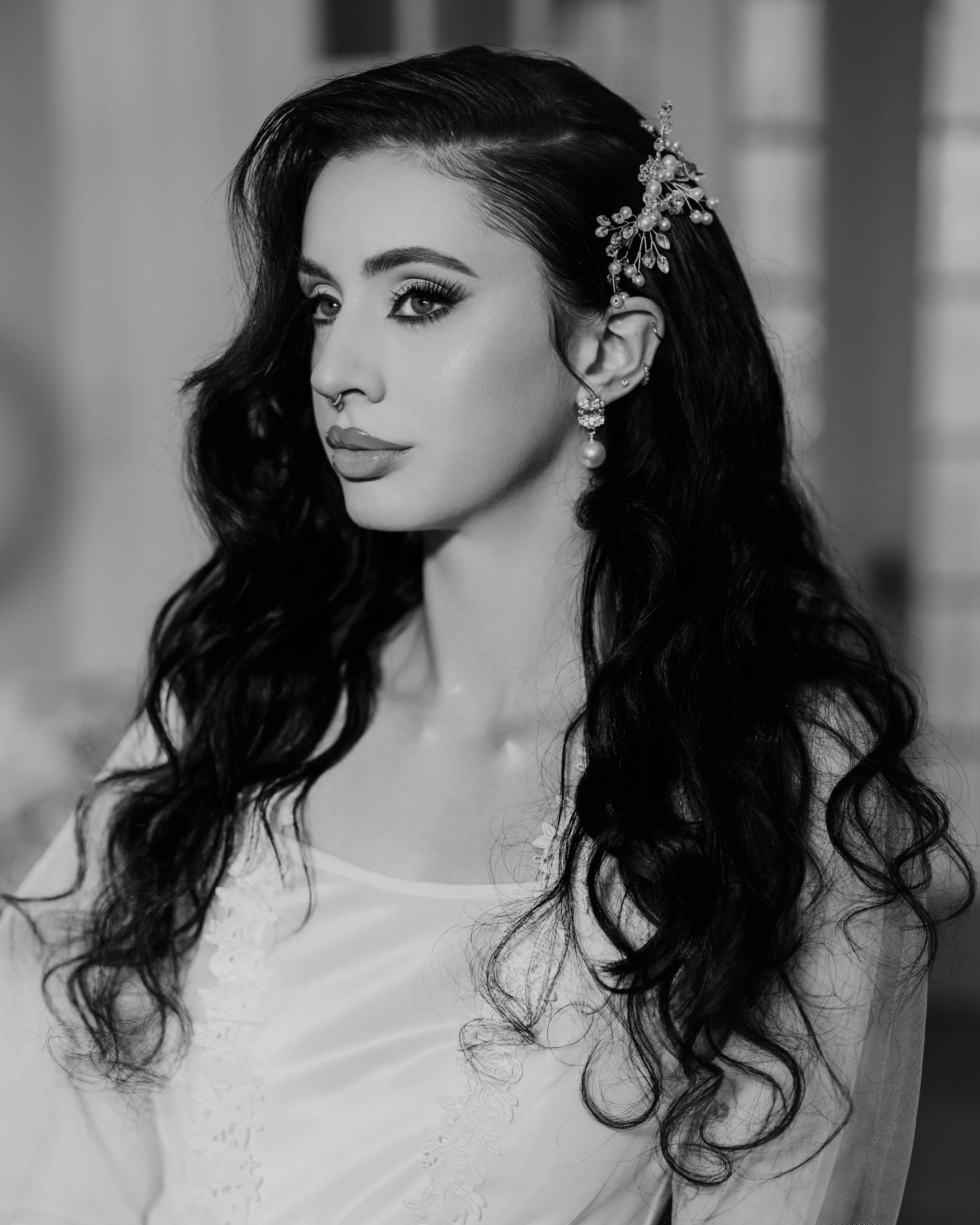 A woman with long wavy black hair wearing jewelry and a white top, with a side profile in a portrait photograph.