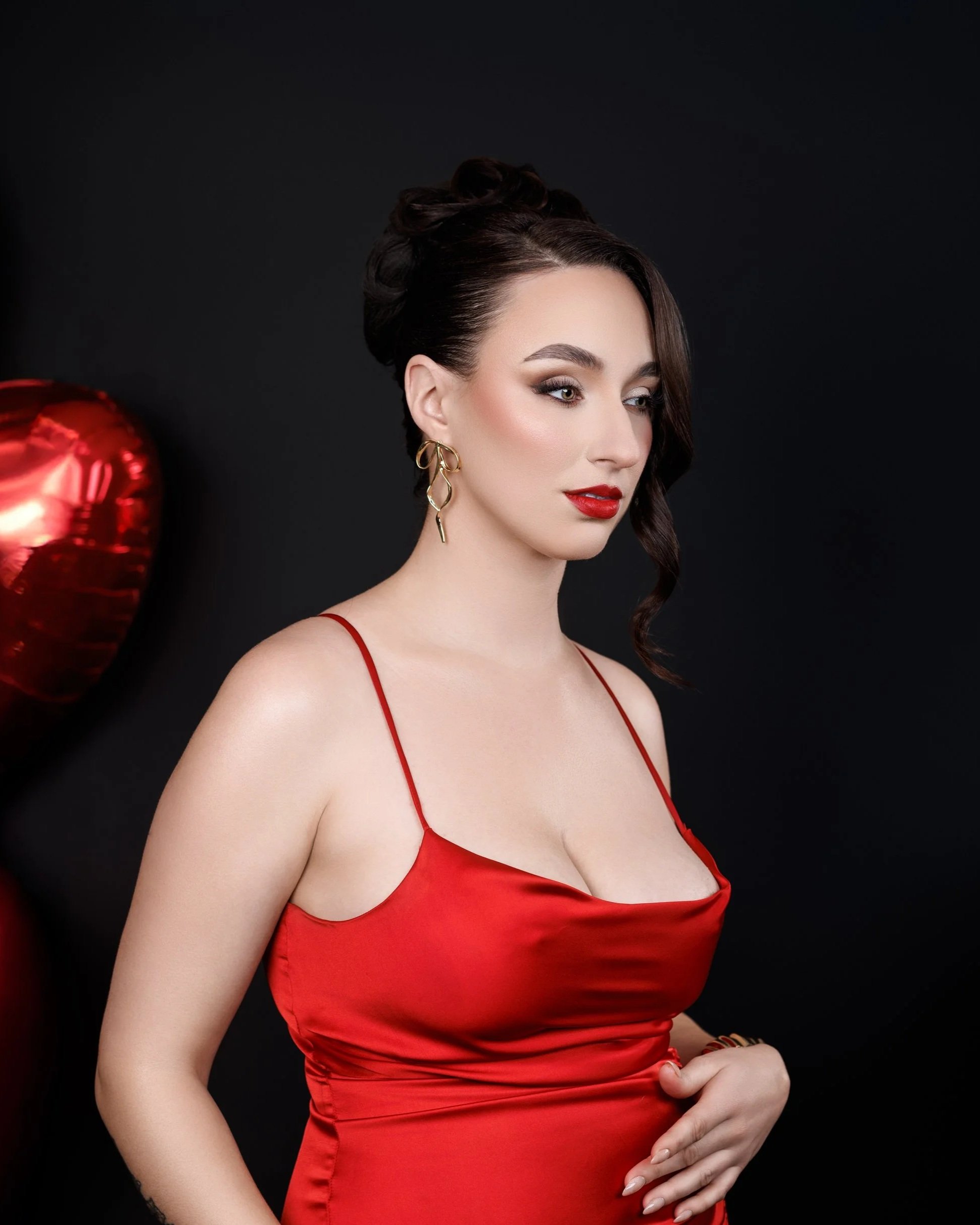 A woman with dark hair in an elegant updo, wearing red lipstick, gold earrings, and a red satin spaghetti strap dress, stands against a black background with a red heart-shaped balloon partially visible on the left.