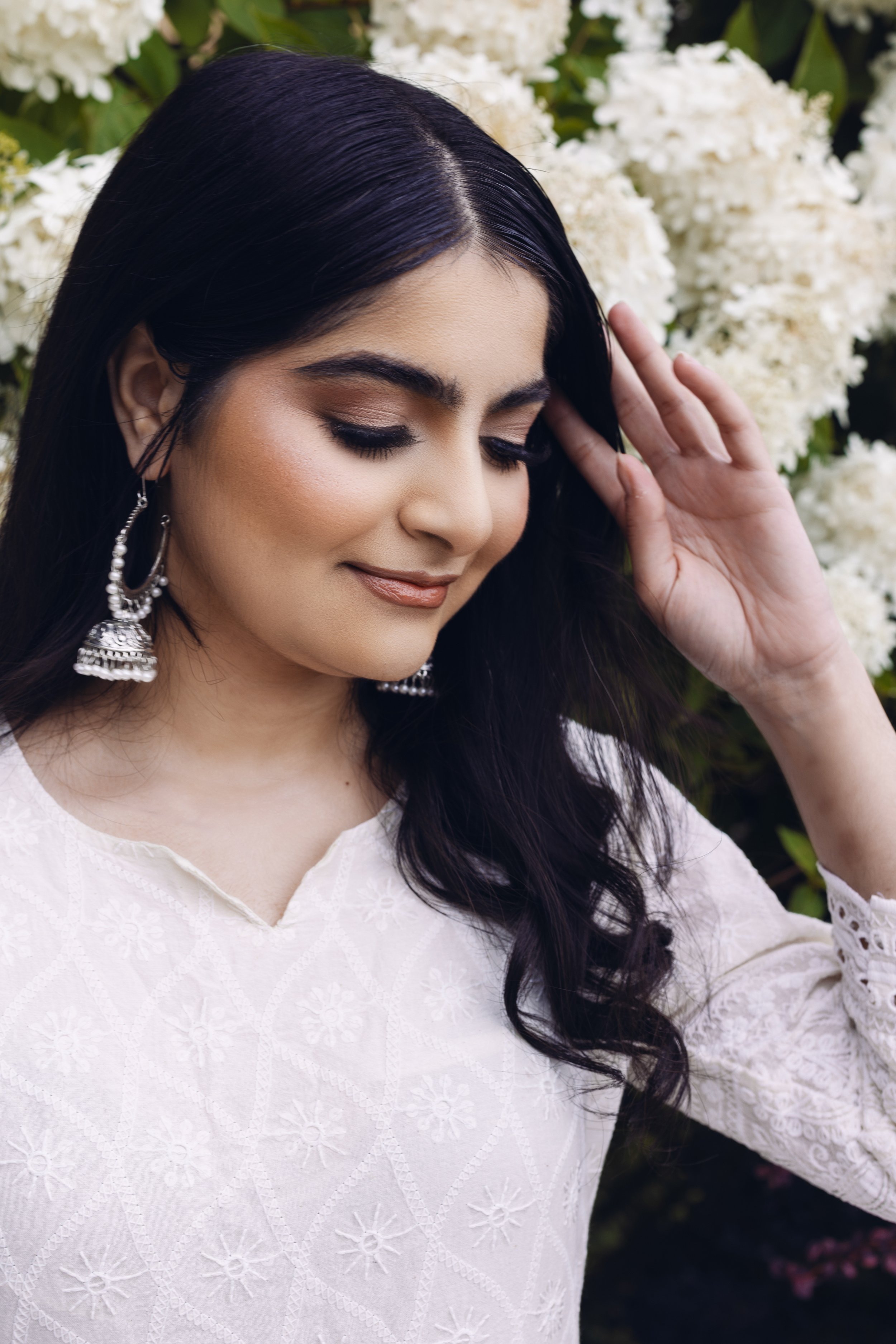 A woman with long black hair, wearing white embroidered clothing and large silver earrings, is standing in front of white flowers, with her eyes closed and her hand lightly touching her head.