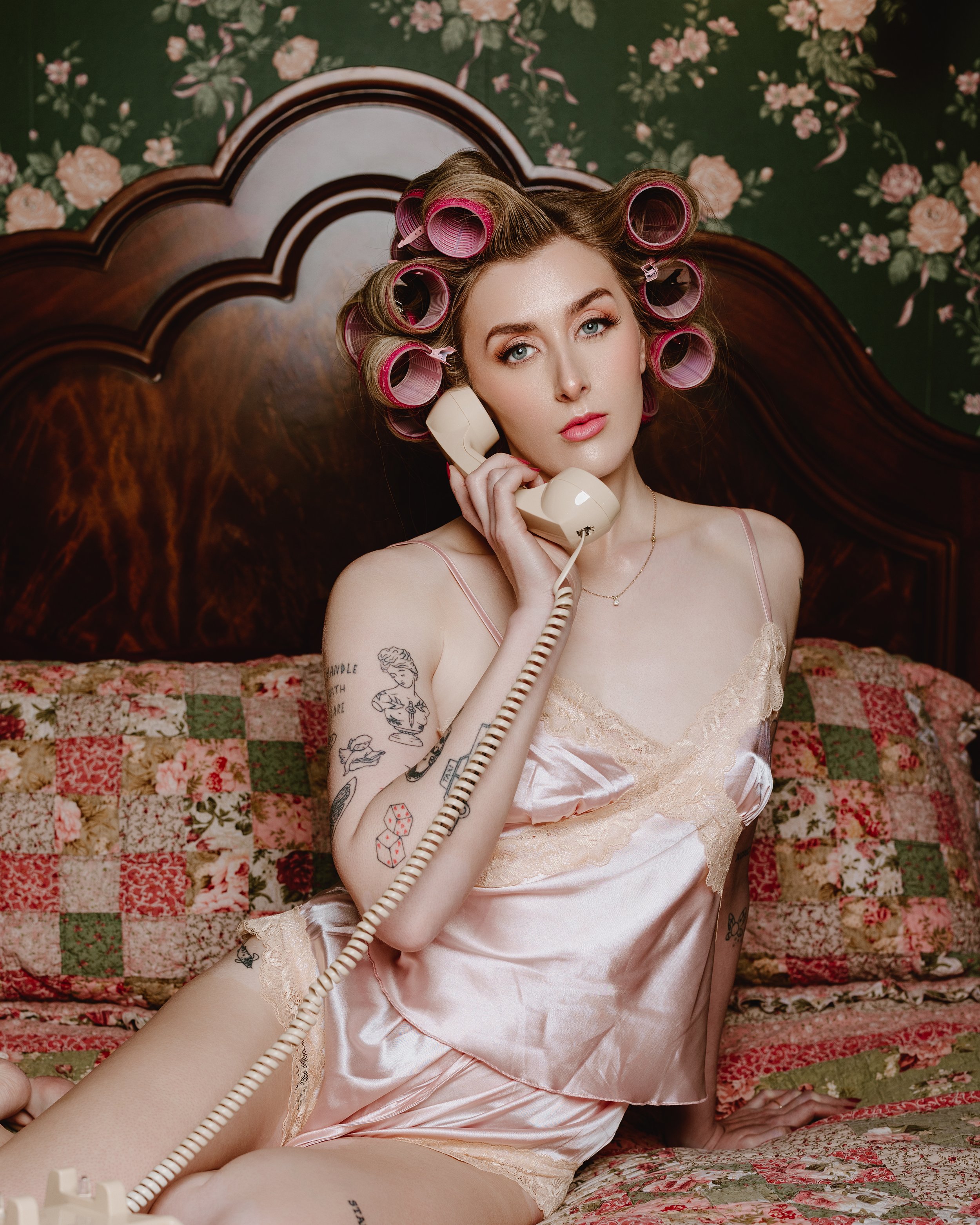 A young woman with pink hair rollers, wearing pink satin pajamas, sitting on a bed with a floral patchwork quilt, talking on a vintage beige landline telephone, with tattoos on her arm and leg, in a bedroom with dark wooden headboard and floral wallpaper.