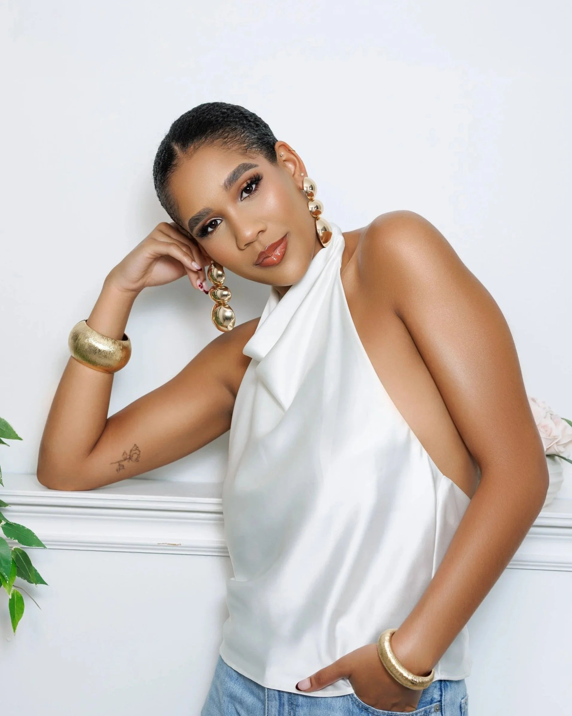 A woman with short dark hair, wearing a white satin top, blue jeans, large gold earrings, a gold bracelet, and a gold bangle, leaning on a white surface against a plain white background.