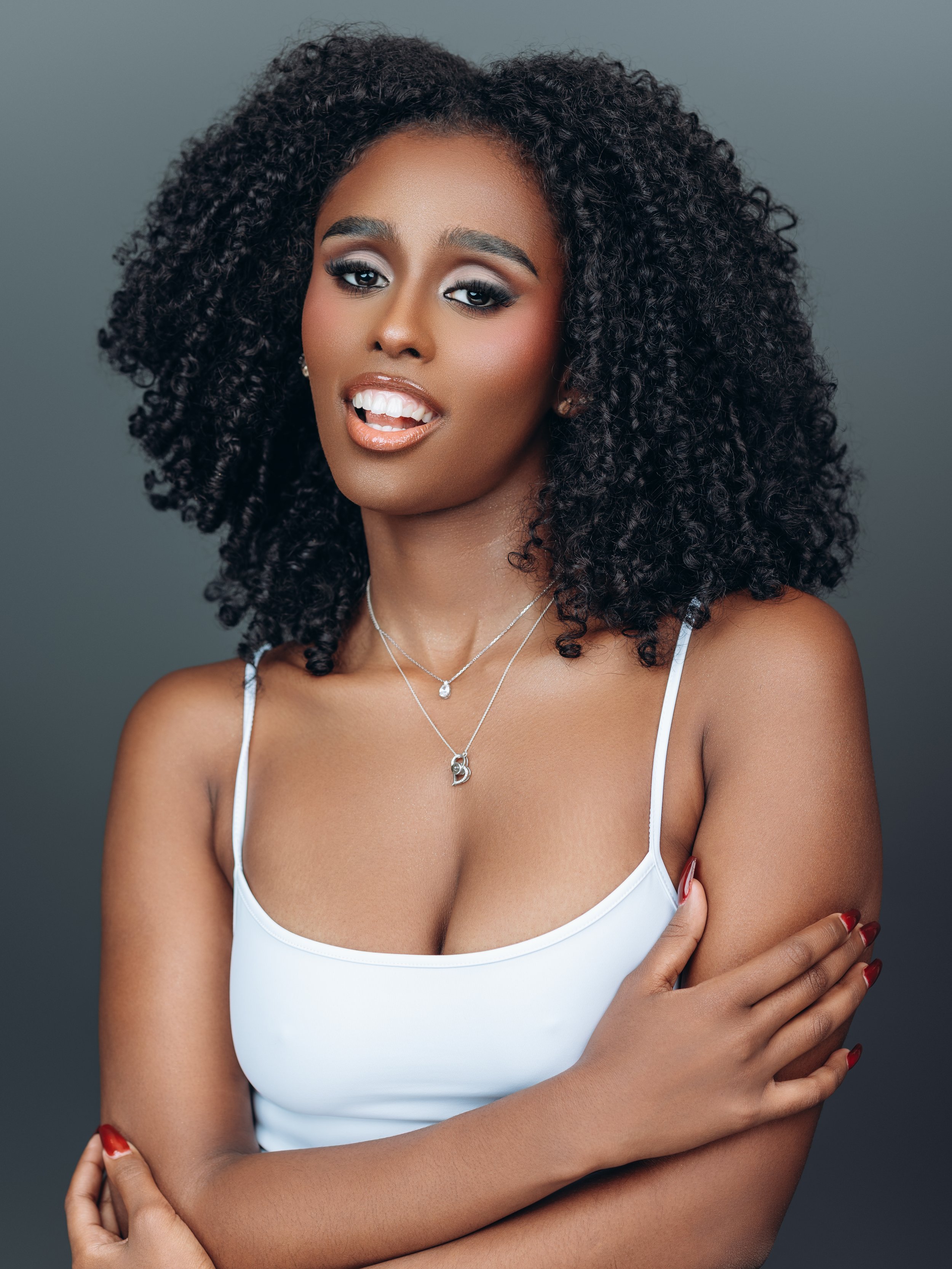 A woman with dark, curly hair wearing a white tank top, jewelry, and makeup, posing against a gray background.