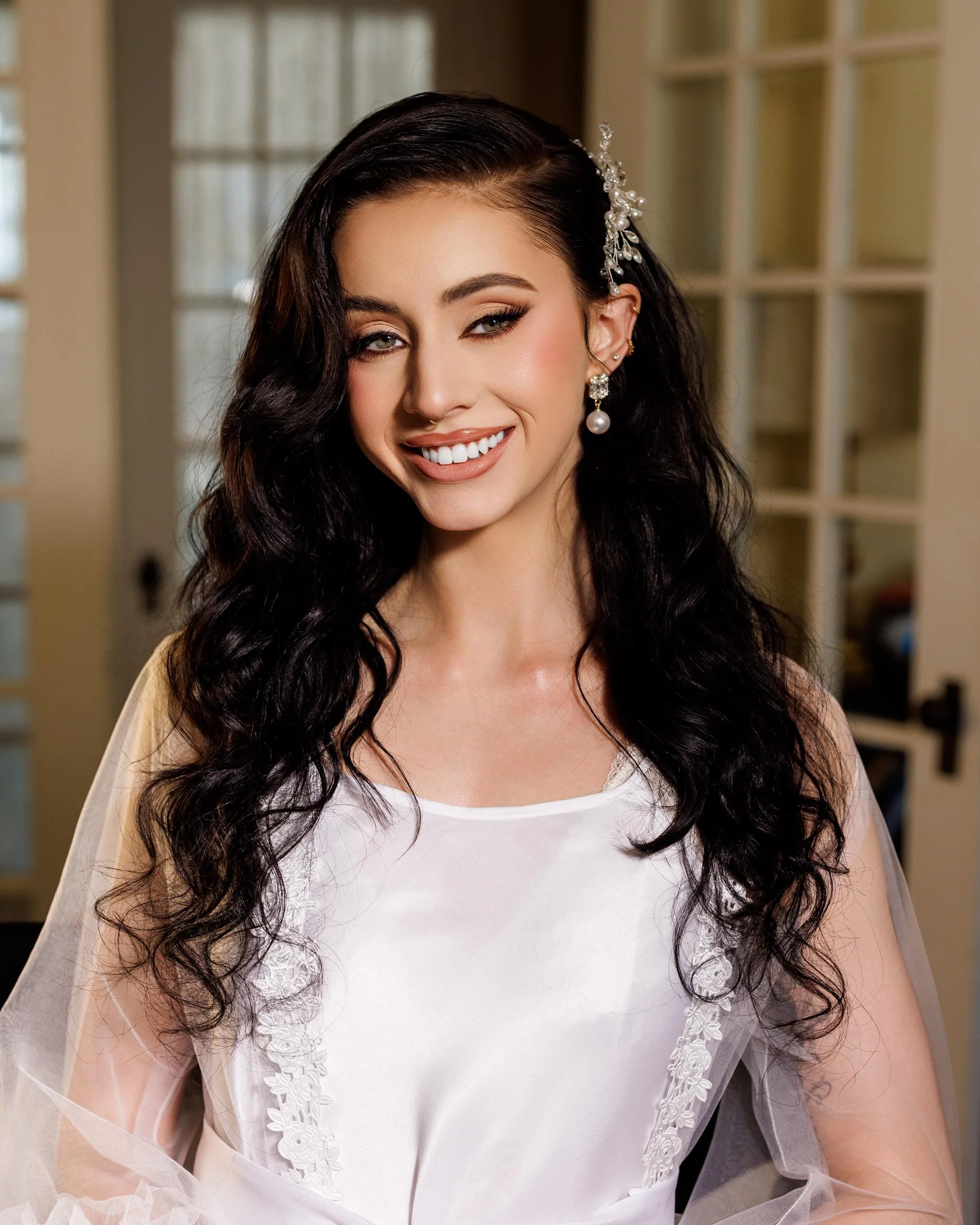 A woman with long dark wavy hair, wearing wedding dress with lace details and pearl earrings, smiling indoors near a window.