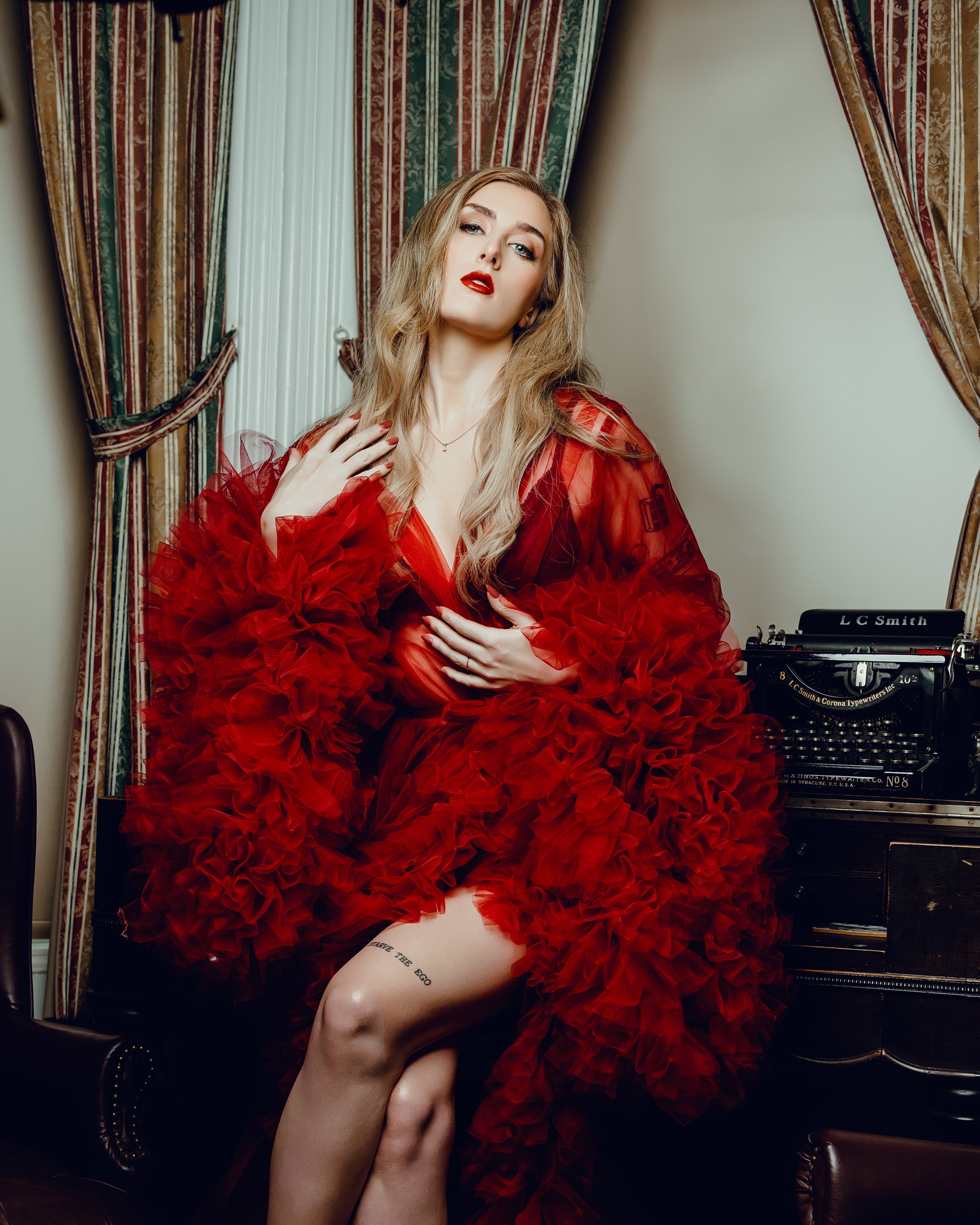 A woman dressed in a red ruffled dress sitting on a chair beside a vintage typewriter.