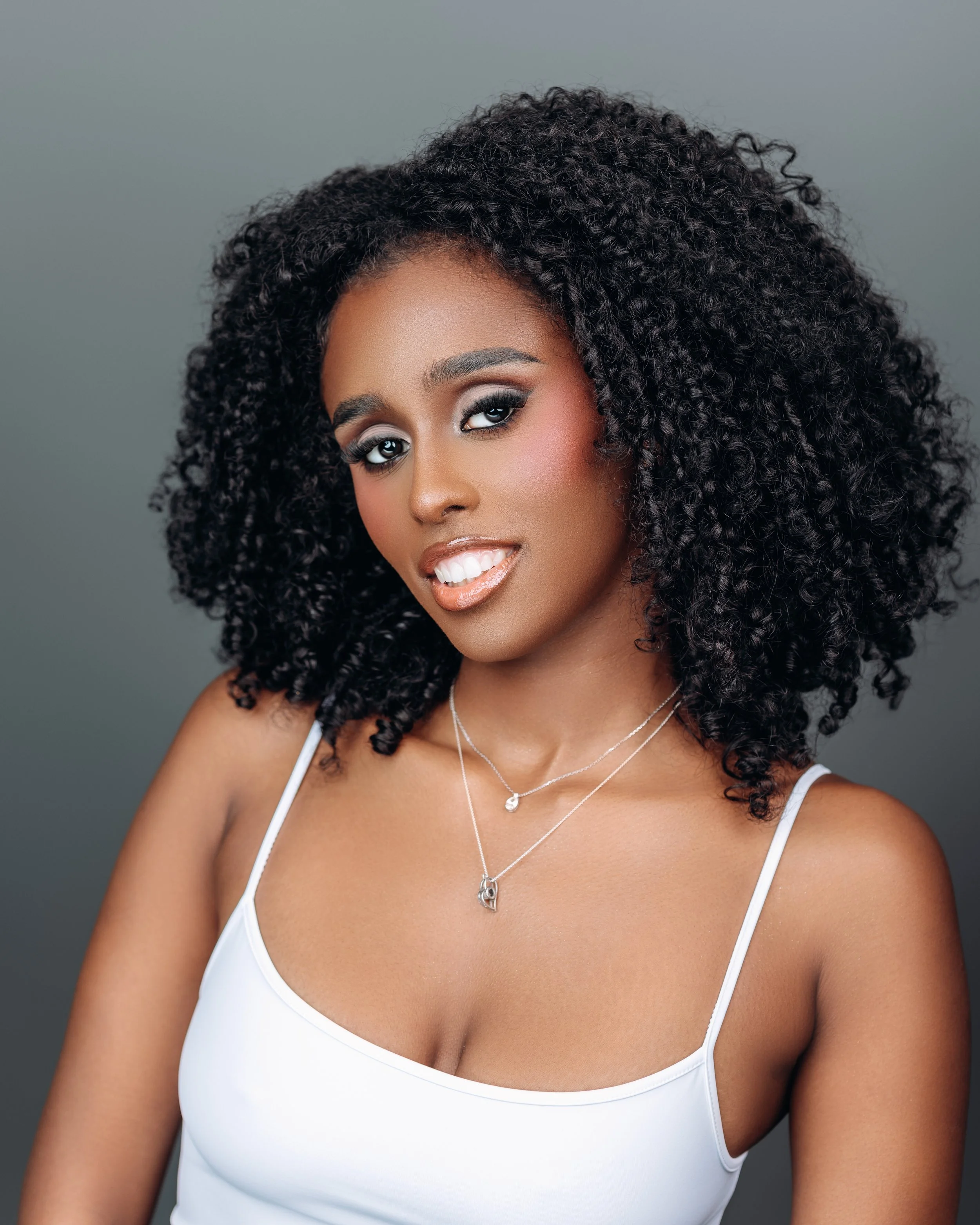 A woman with curly black hair, wearing a white tank top and layered necklaces, smiling against a gray background.