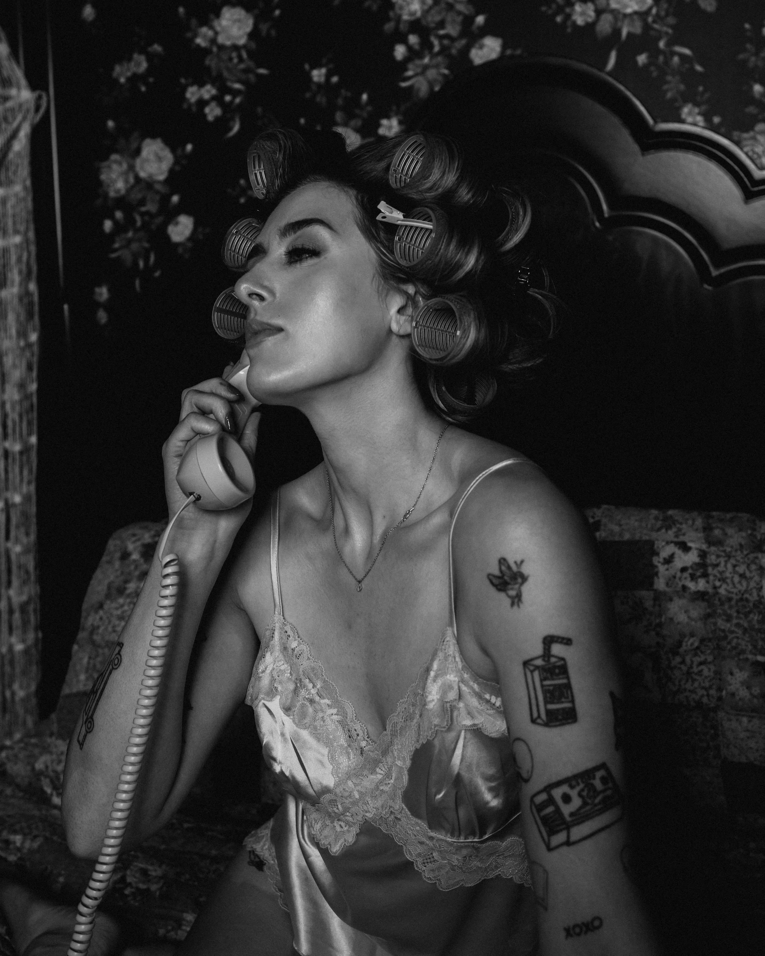 A woman with curlers in her hair, wearing a satin camisole, talks on a vintage rotary phone.