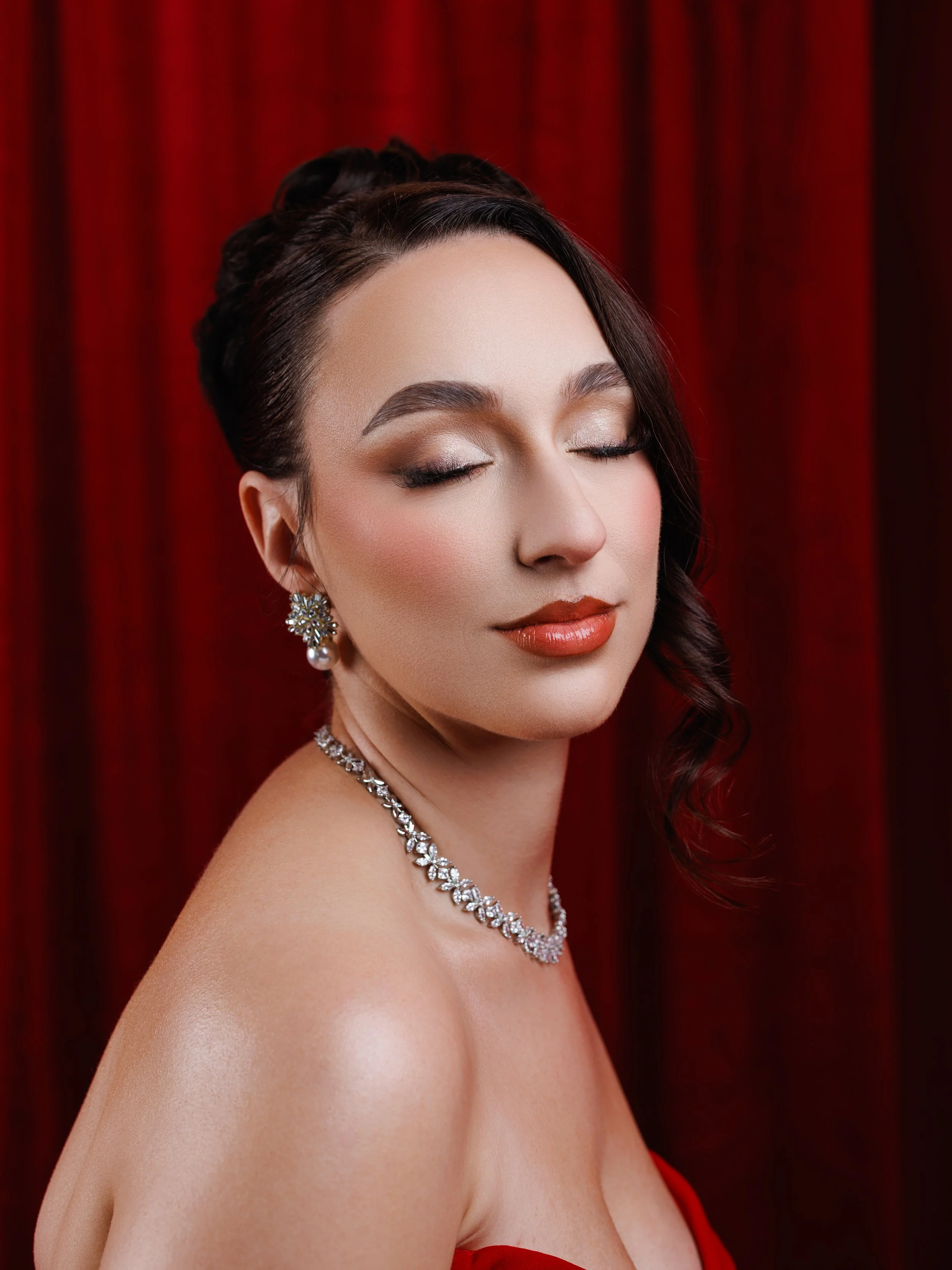 A woman with dark hair styled in soft curls, closed eyes, wearing diamond jewelry, and a red strapless dress against a red curtain background.