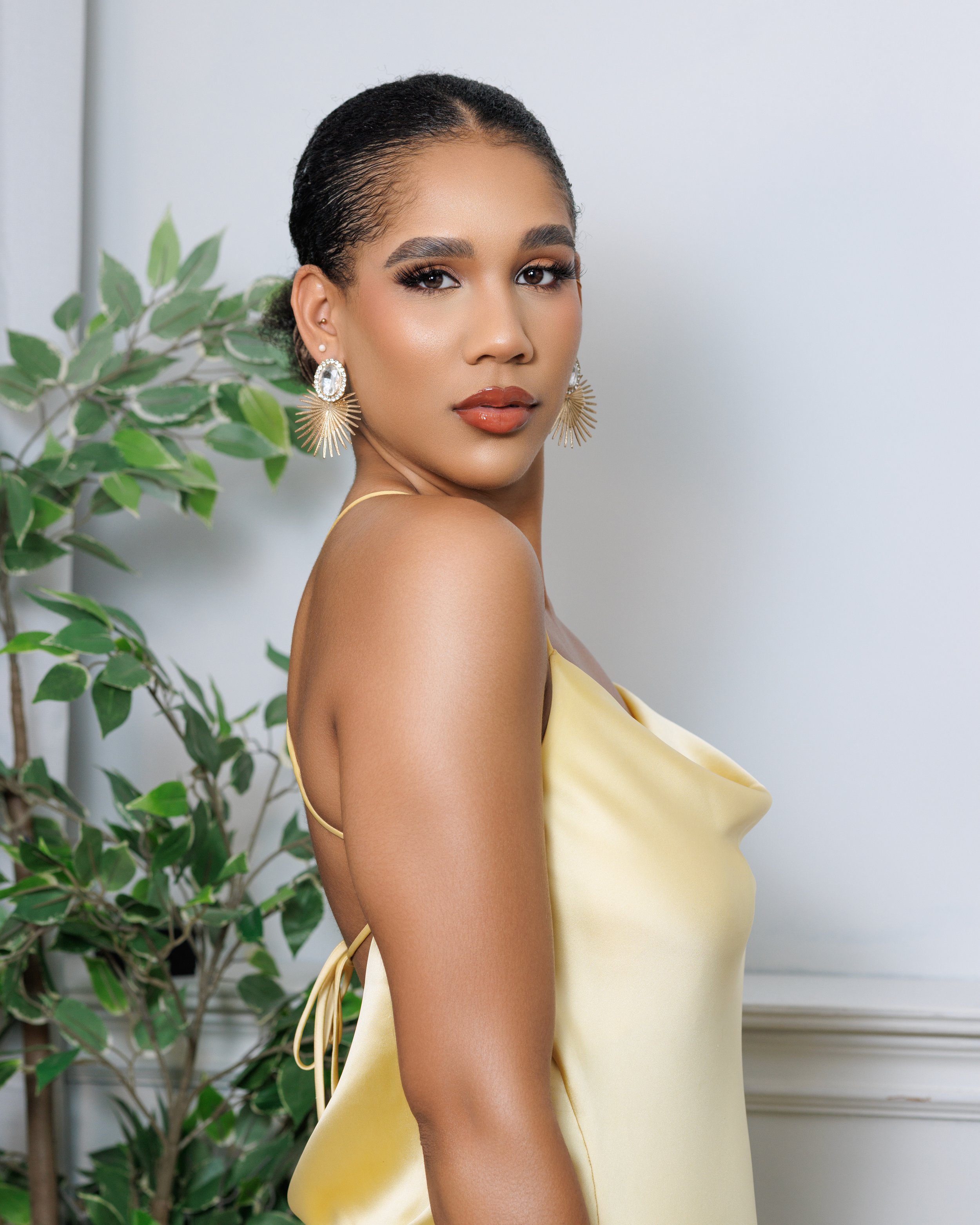 A woman with medium brown skin, dark styled hair, and makeup, wearing gold earrings and a yellow satin dress, standing indoors near a white wall and green plant.