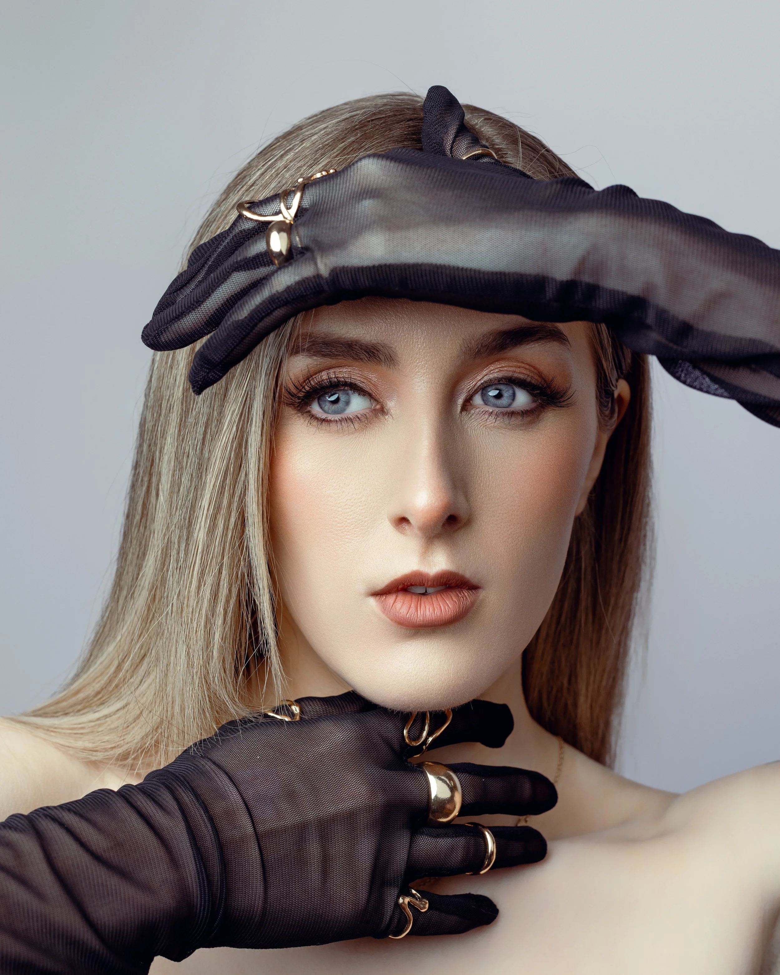 A close-up of a woman with blue eyes and makeup, wearing sheer black gloves with gold rings, posing with her hands near her face against a plain background.