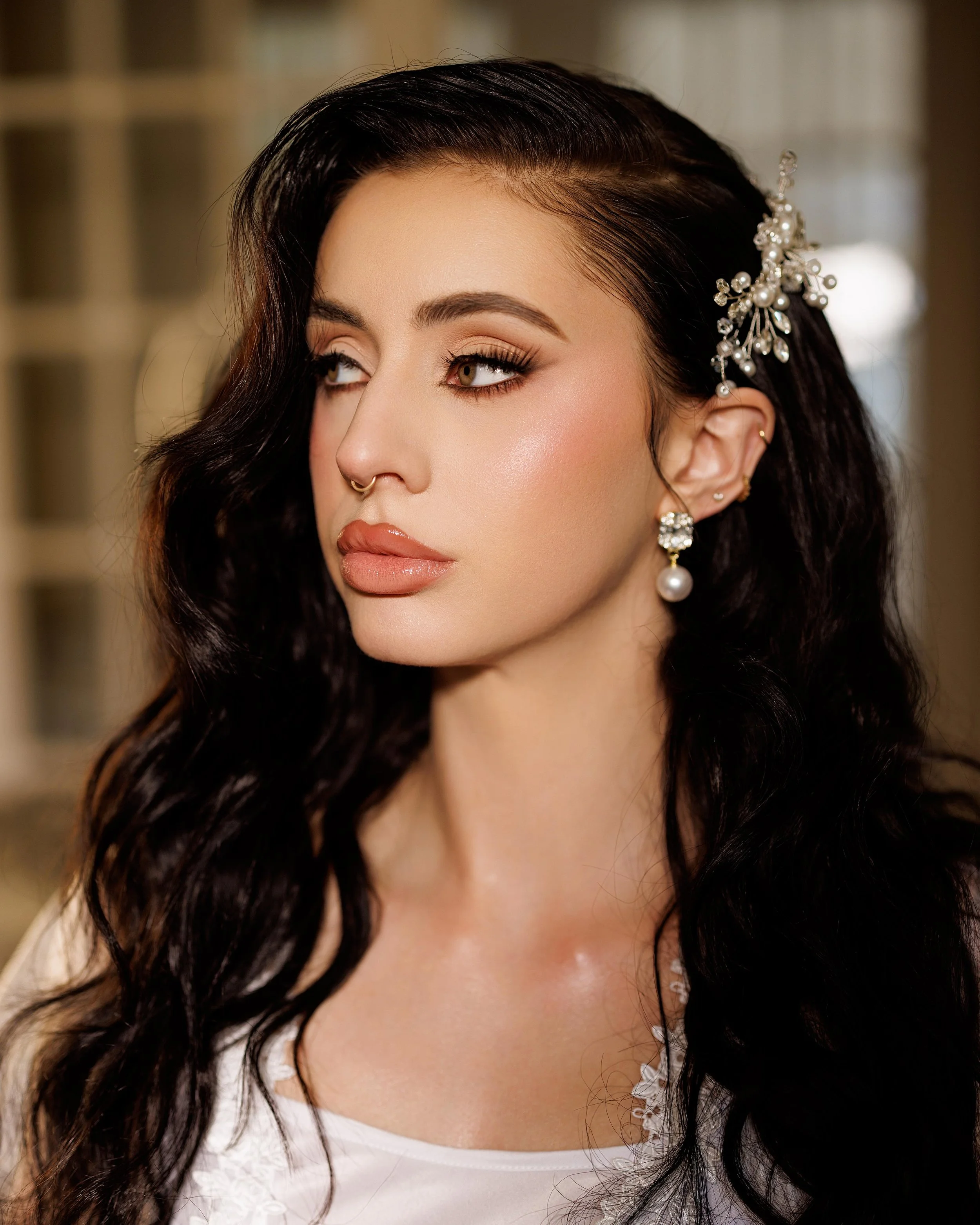 Close-up portrait of a woman with dark wavy hair, wearing jewelry, makeup, and a white top, indoors.