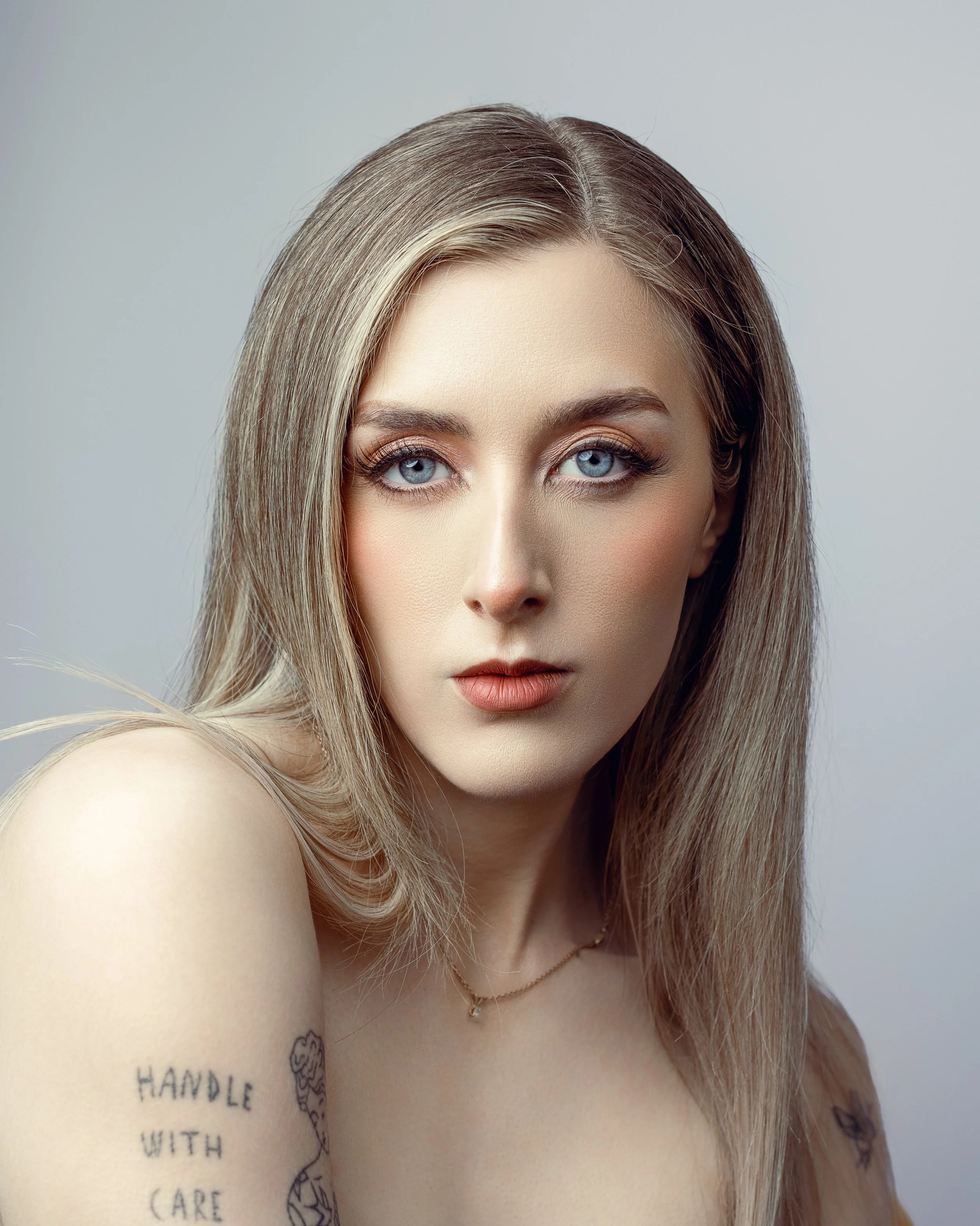 Close-up portrait of a young woman with blue eyes and blonde hair, wearing a delicate necklace, with visible tattoos on her shoulder.
