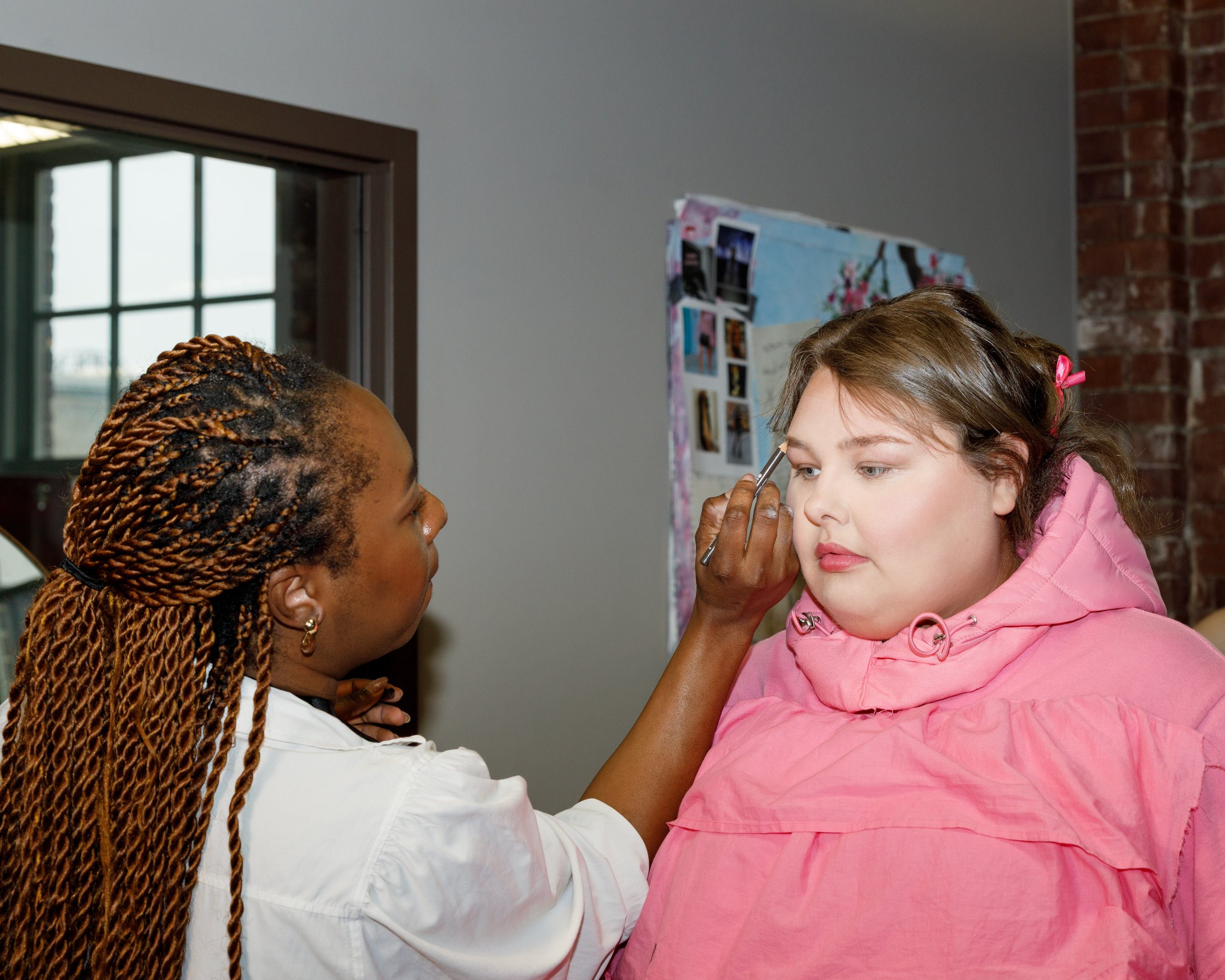 Makeup artist applying makeup to a young woman in a pink hoodie.