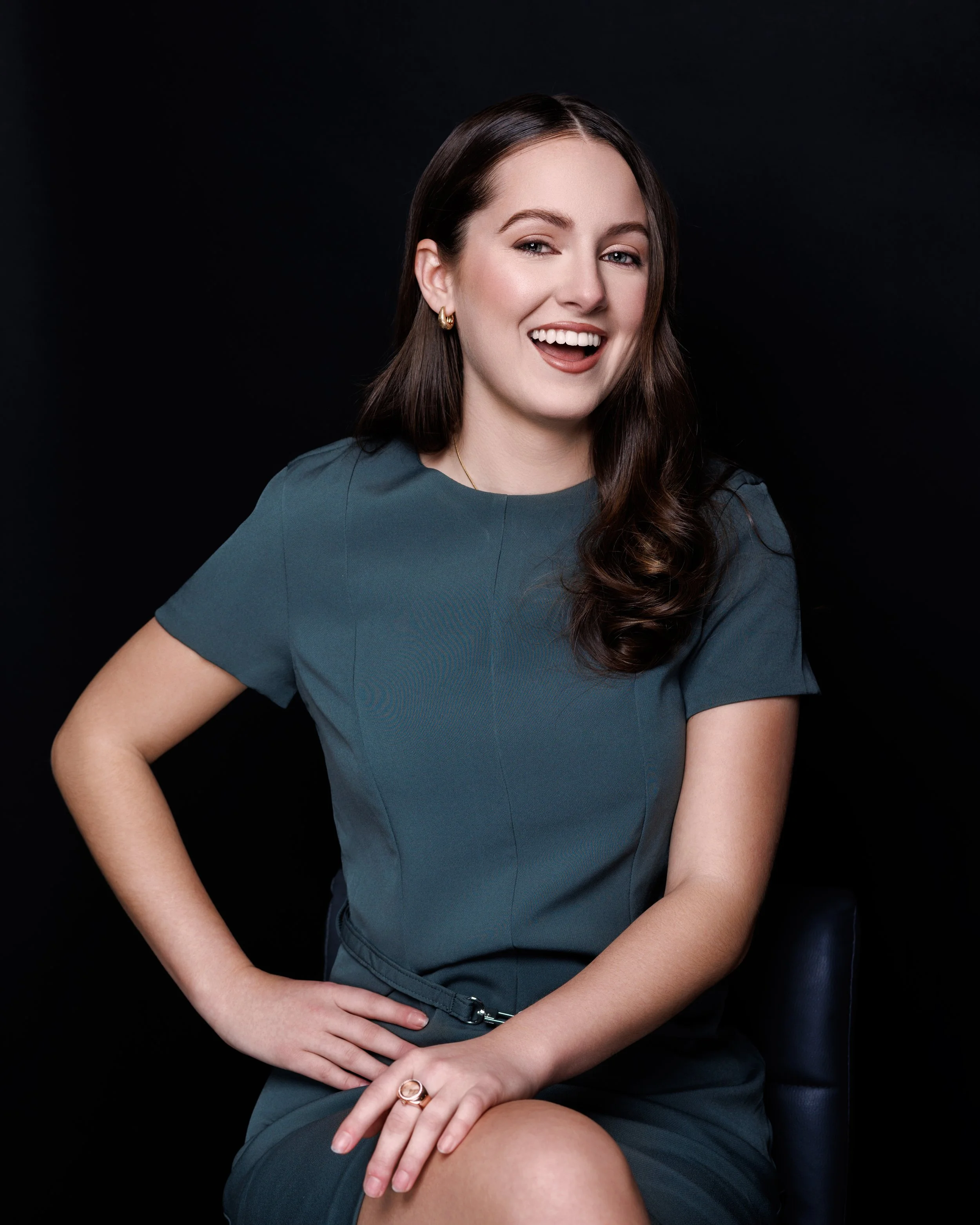A woman with long brown hair winks and smiles, wearing a blue dress, sitting against a black background.