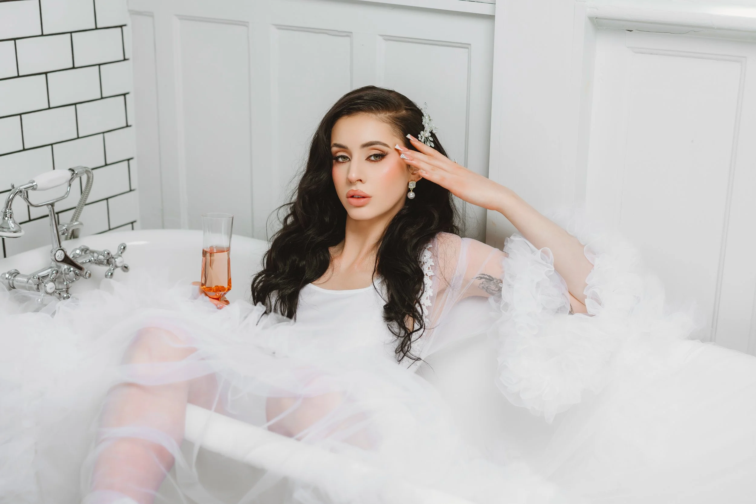 A woman with long dark hair, makeup, and earrings, relaxing in a bathtub with white ruffled fabric, a glass of pink drink on the edge, in a bathroom with white paneling and brick wall.