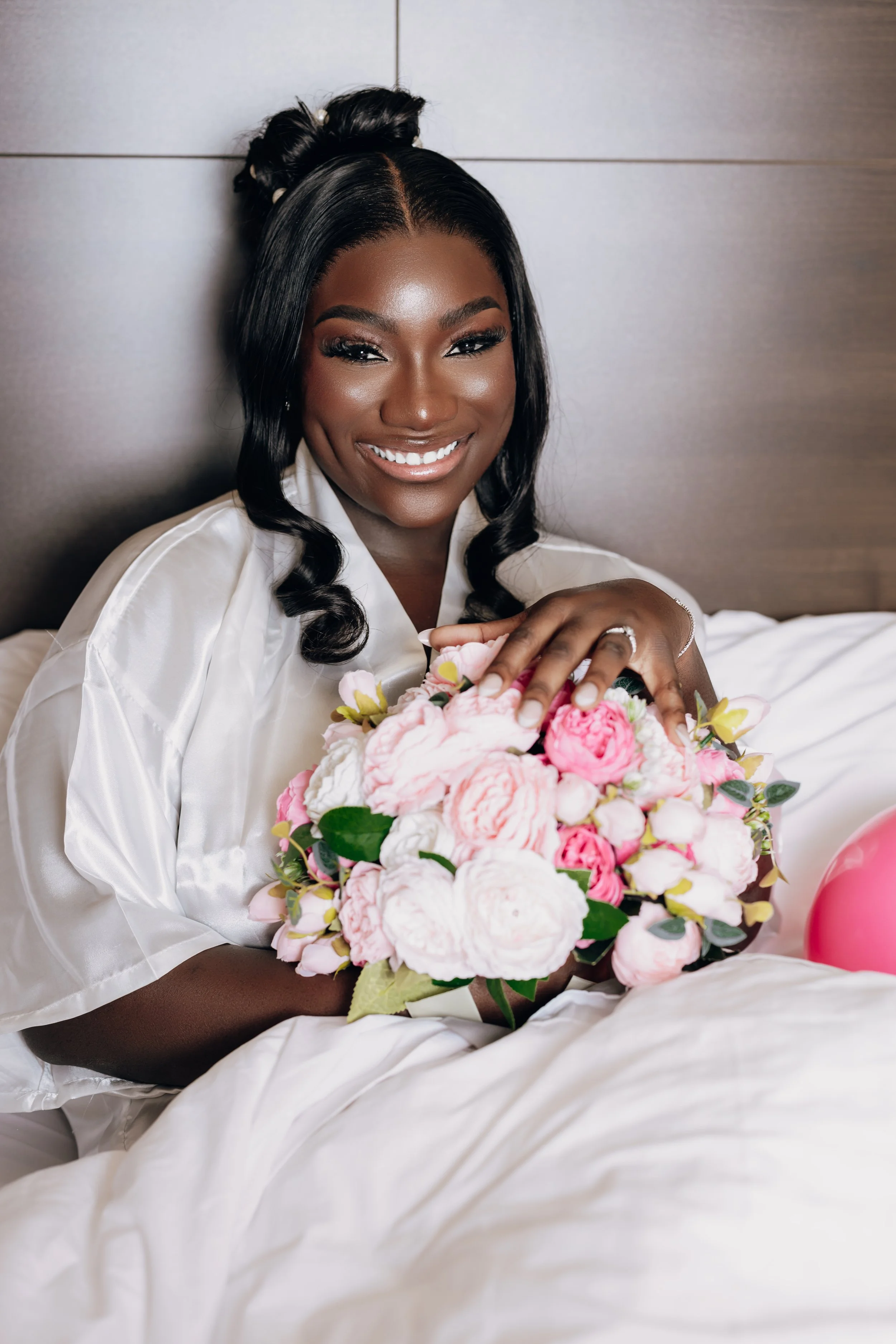 A woman dressed in satin pajamas smiling while holding a bouquet of pink and white flowers on a bed.