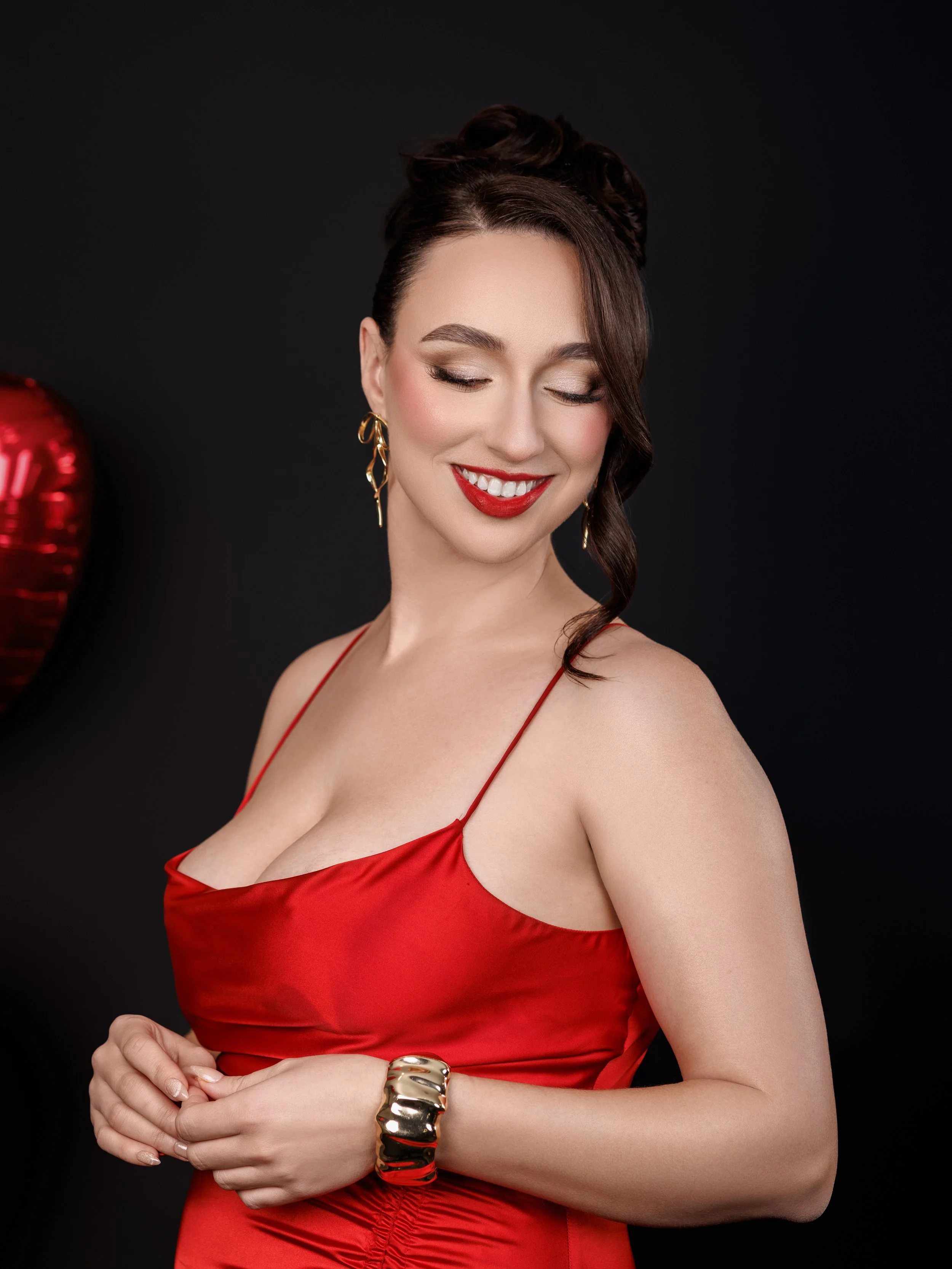 A woman wearing a red satin dress with spaghetti straps, gold earrings, and a gold bracelet, smiling with her eyes closed, set against a dark background with a red heart-shaped balloon on the left side.