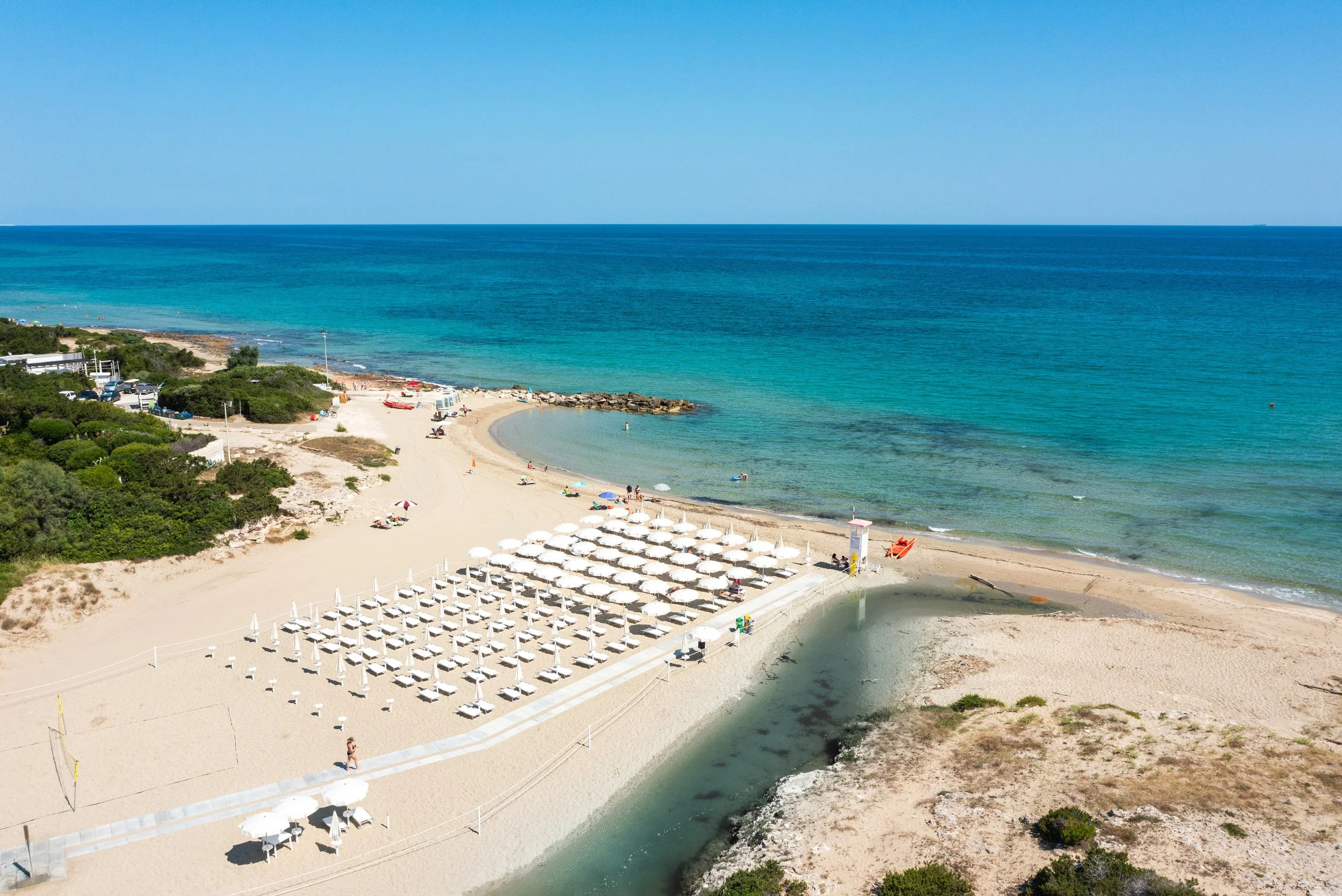 A sandy beach with rows of white umbrellas and lounge chairs, a small river flowing into the ocean, green bushes, and a clear blue sky with calm ocean waters in the background.