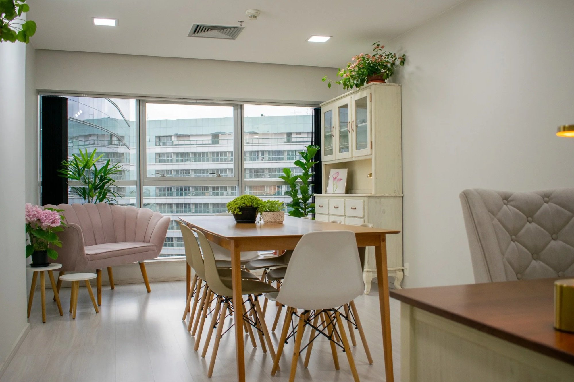 Sala de jantar iluminada com mesa de madeira, 6 cadeiras brancas, poltrona rosa, estante branca com plantas e uma janela grande com vista para edifícios