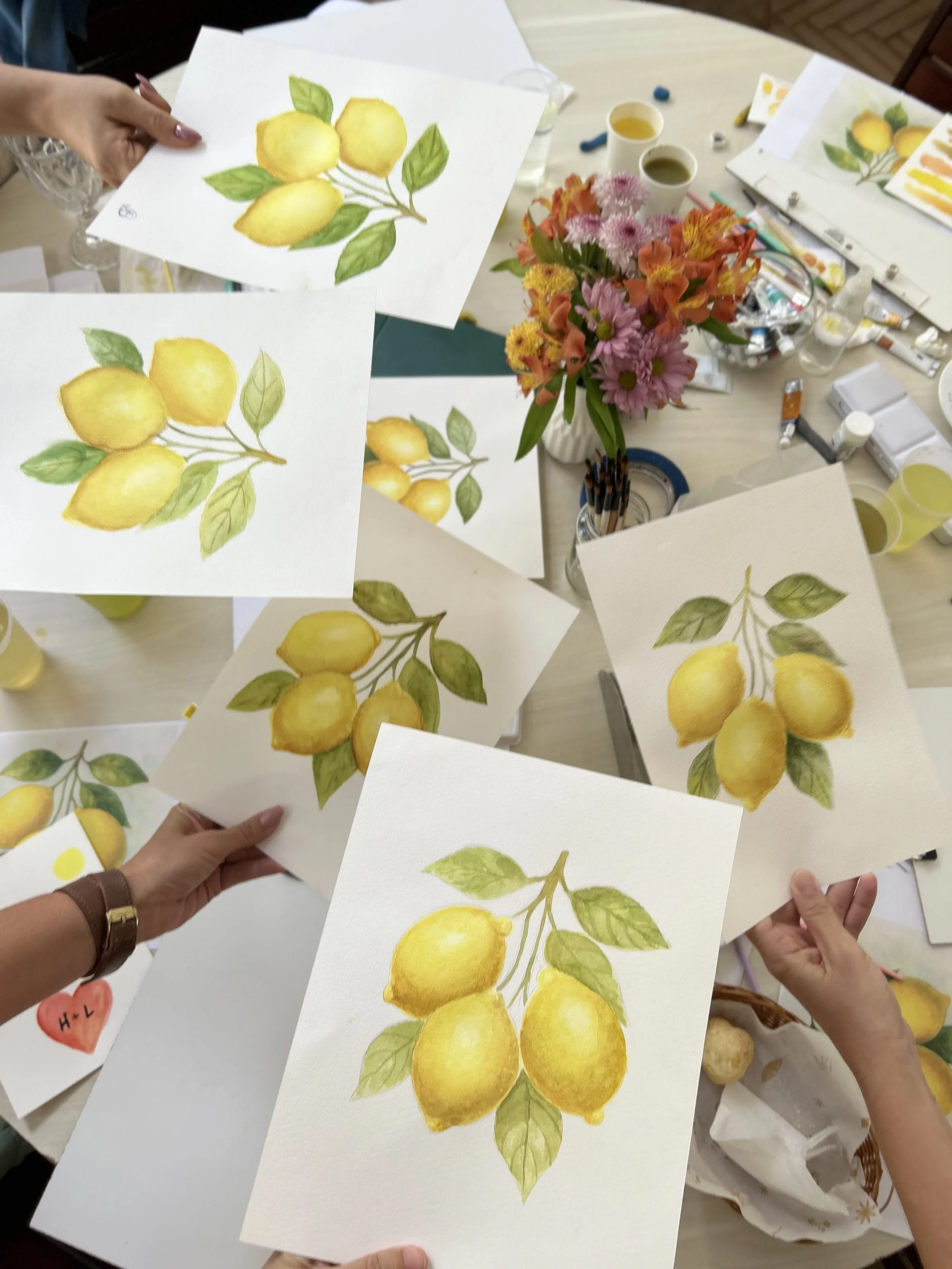 Pessoas segurando desenhos de limões pintados à mão, com uma mesa com tintas, pincéis e arranjo de flores ao fundo.