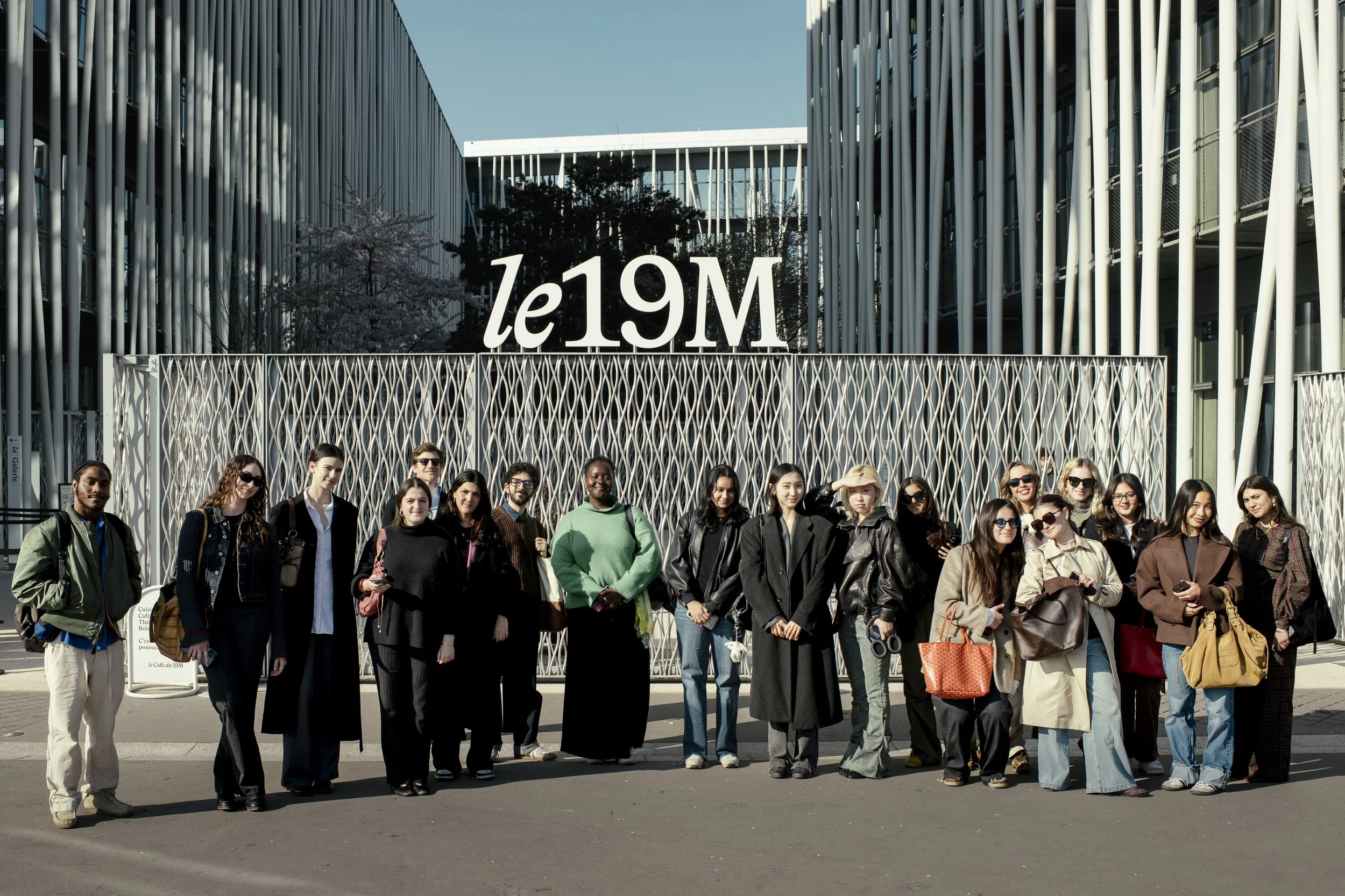 A diverse group of people standing in front of a modern building with a sign that reads 'le19M' above the gate.