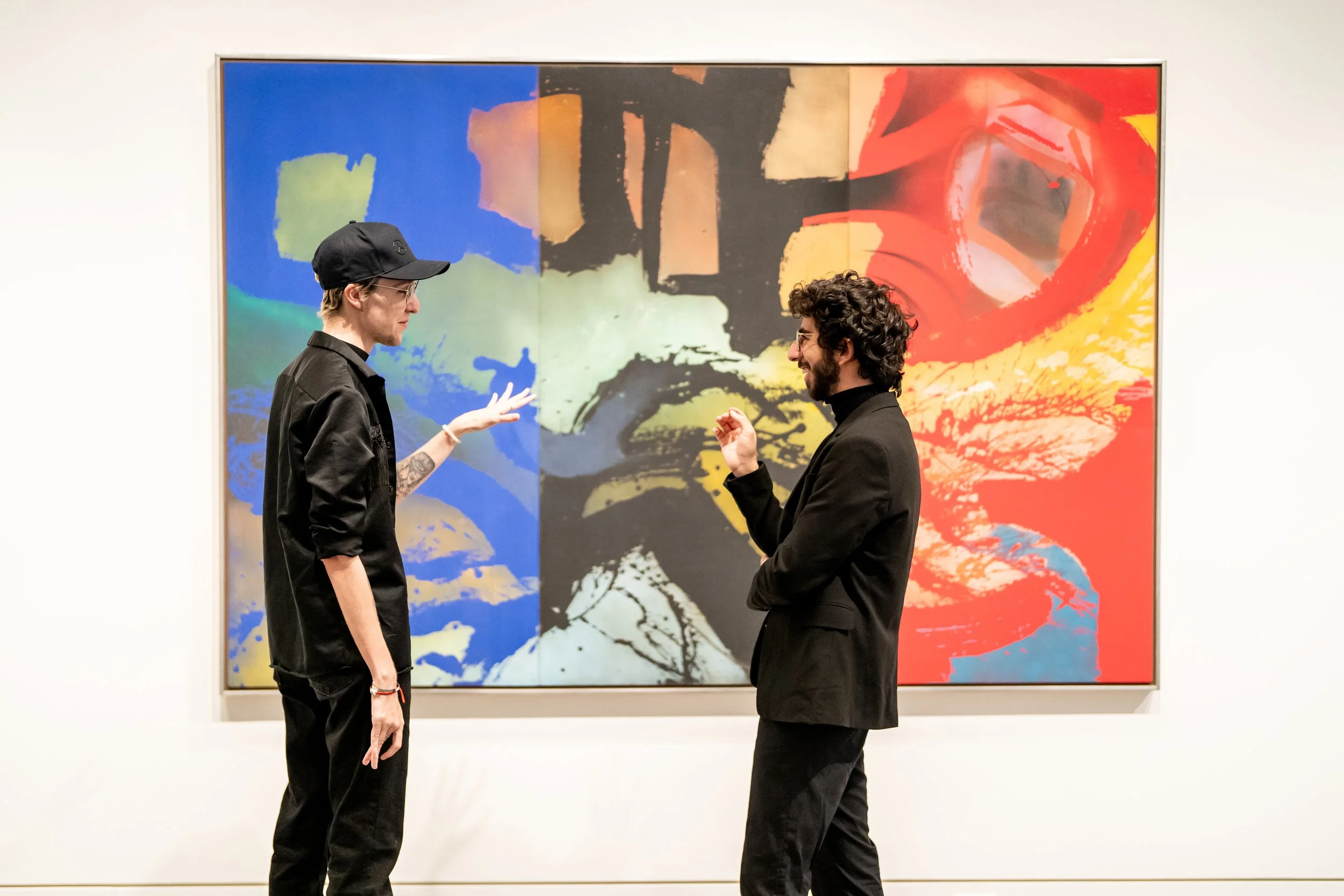 Two men engage in conversation in front of a colorful abstract painting at an art gallery. The man on the left wears a black cap, black jacket, and glasses, with tattoos on his arm. The man on the right has curly hair, a beard, and wears a black blazer.