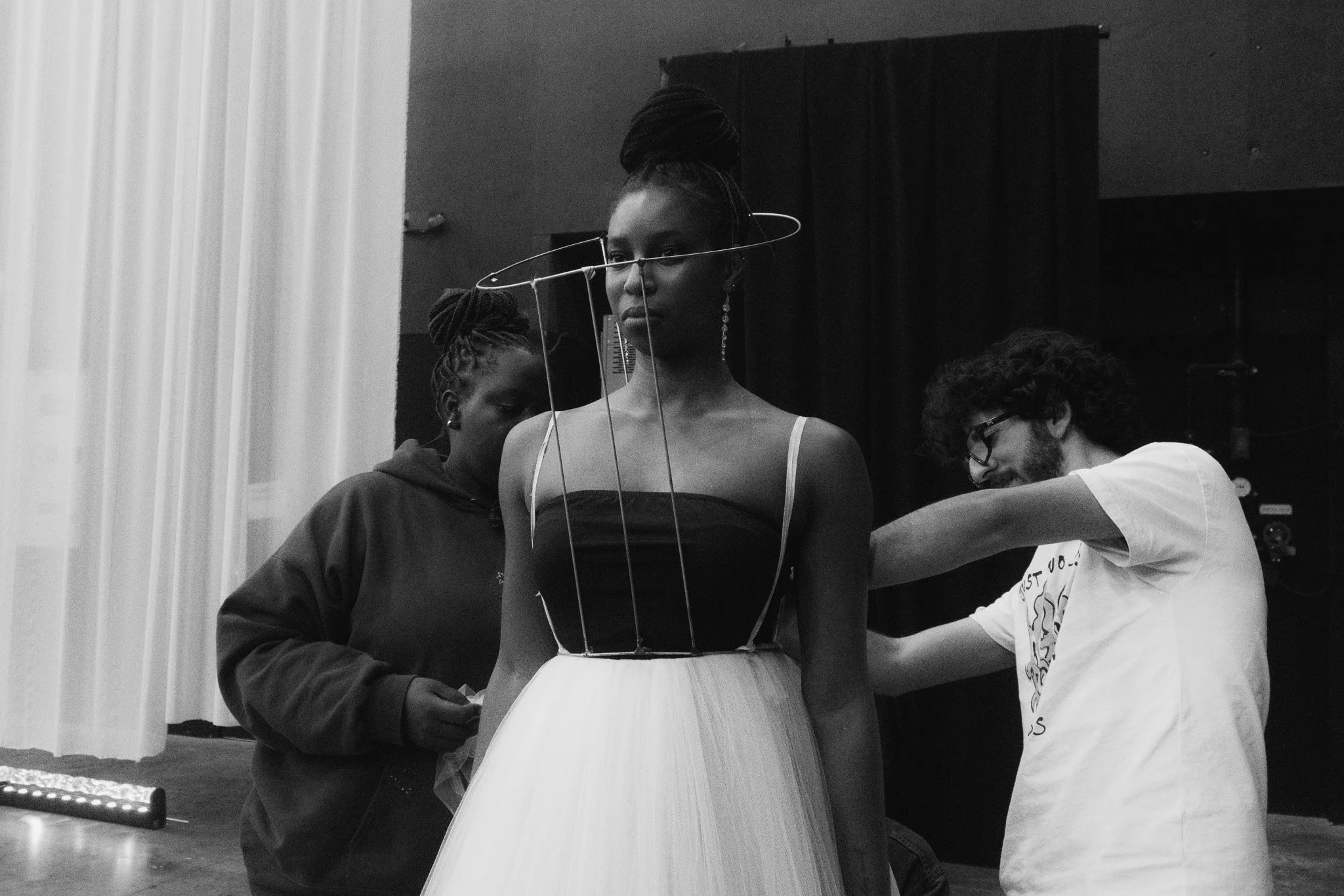 Three people, one woman in a black dress and white tulle skirt, with a wire frame around her head, being fitted for a dress by two assistants in a dressing room.