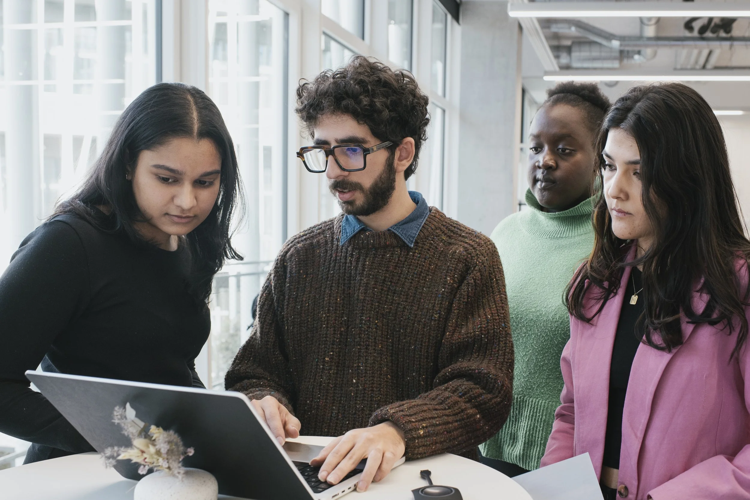 Four young adults gathered around a laptop in a bright office space, looking at the screen and discussing.