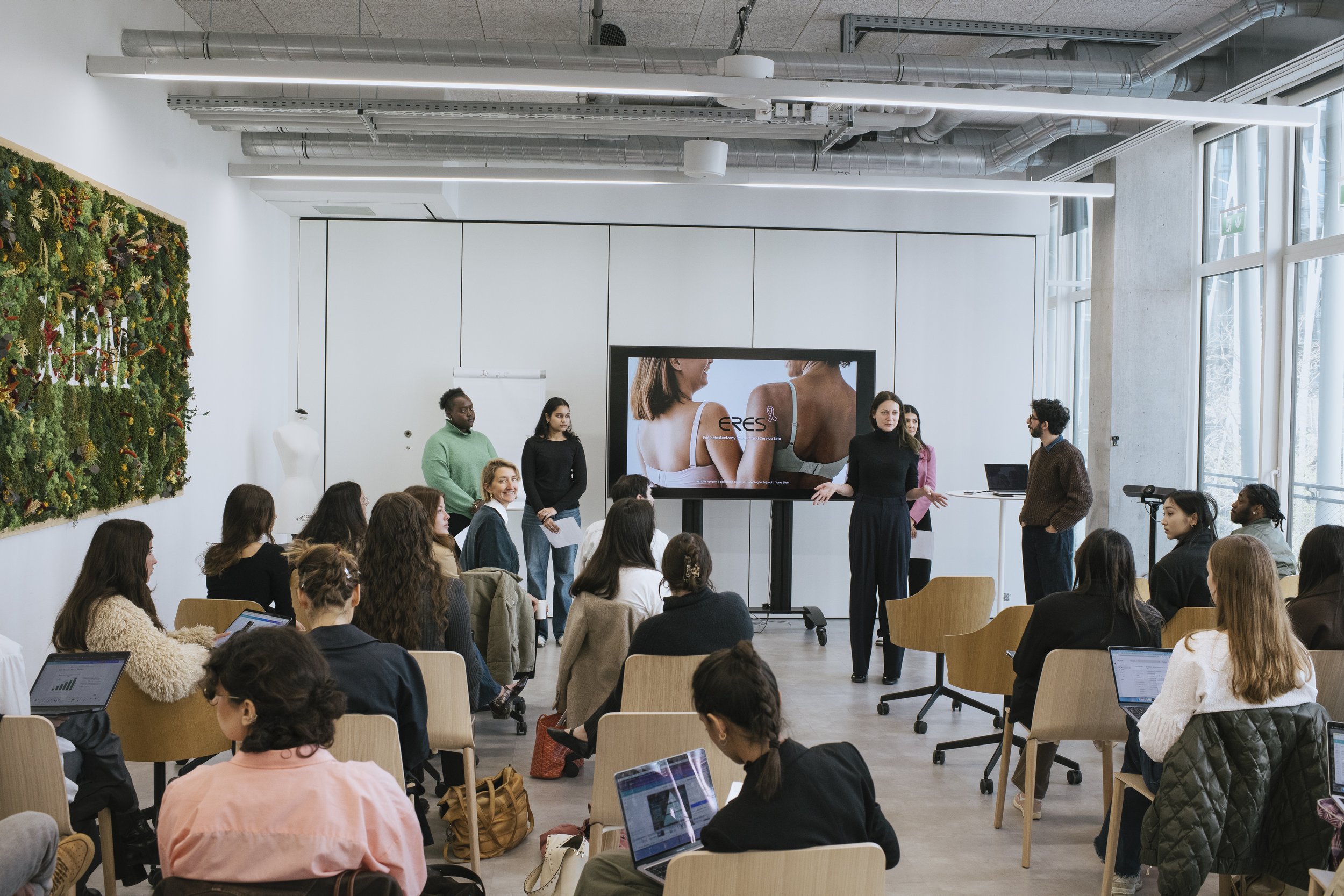 A group of people attending a presentation in a modern conference room. A woman is speaking near a large screen displaying the word "ERES" and an image of two women in lingerie. Several attendees are using laptops and taking notes.