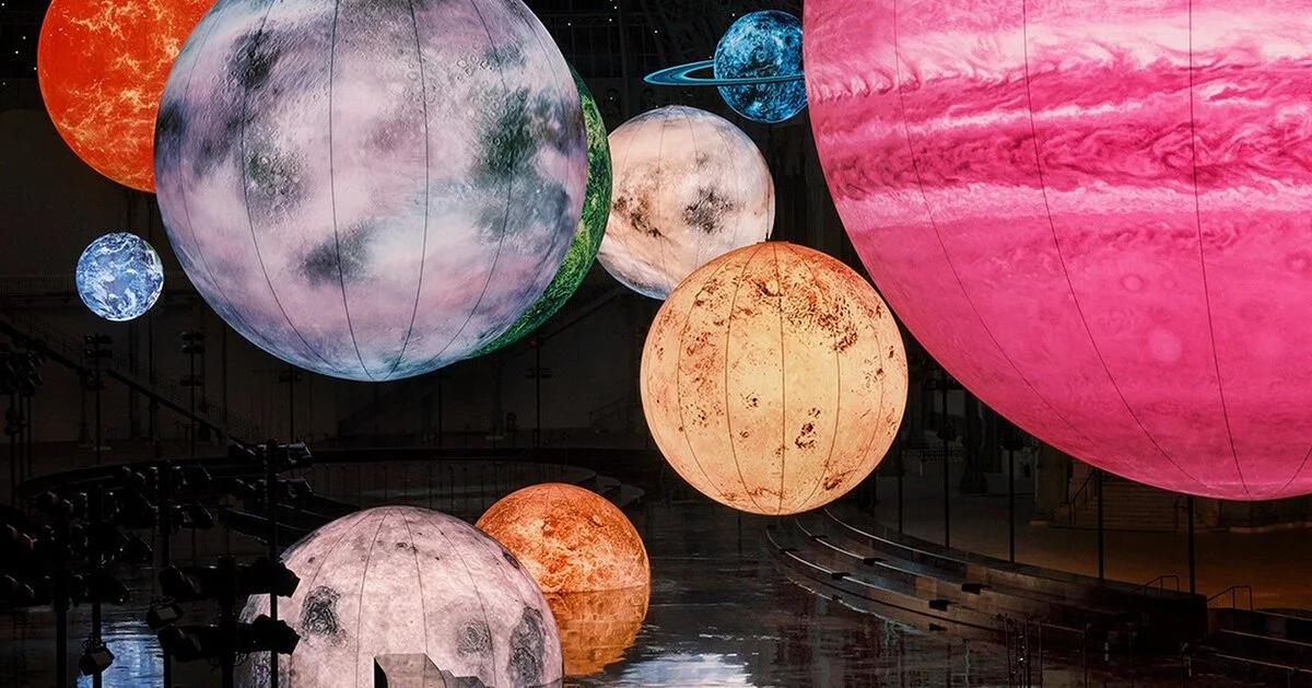 Multiple illuminated paper lanterns designed to look like planets and moons in outer space, hanging indoors.