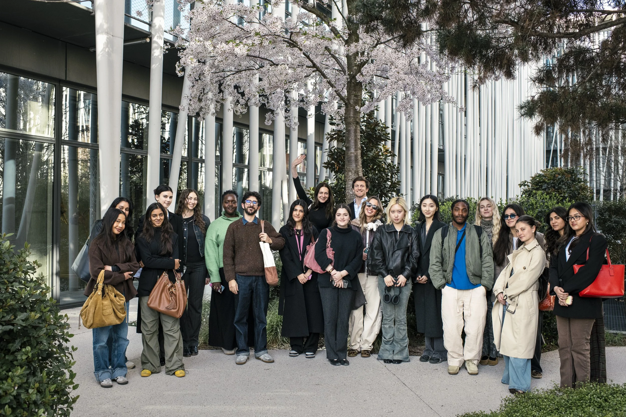 A diverse group of people standing outside in front of modern glass building with trees, some women are holding bags, one person is holding binoculars, and others are smiling and posing for the photo.
