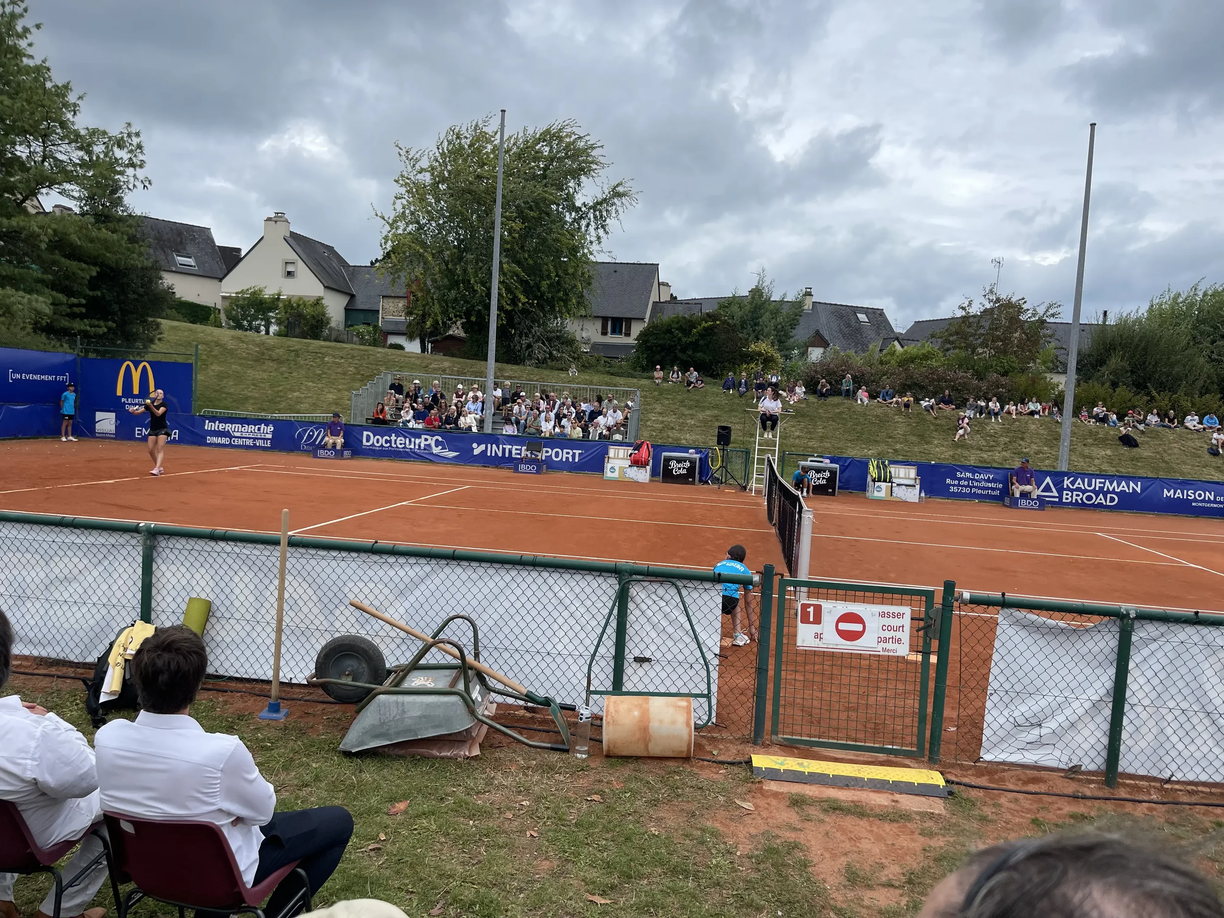Capture d'un match de tennis sur un court en terre battue avec des spectateurs assis au fond et devant des personnes assises sur des chaises, un panneau d'interdiction et des équipements autour du court.