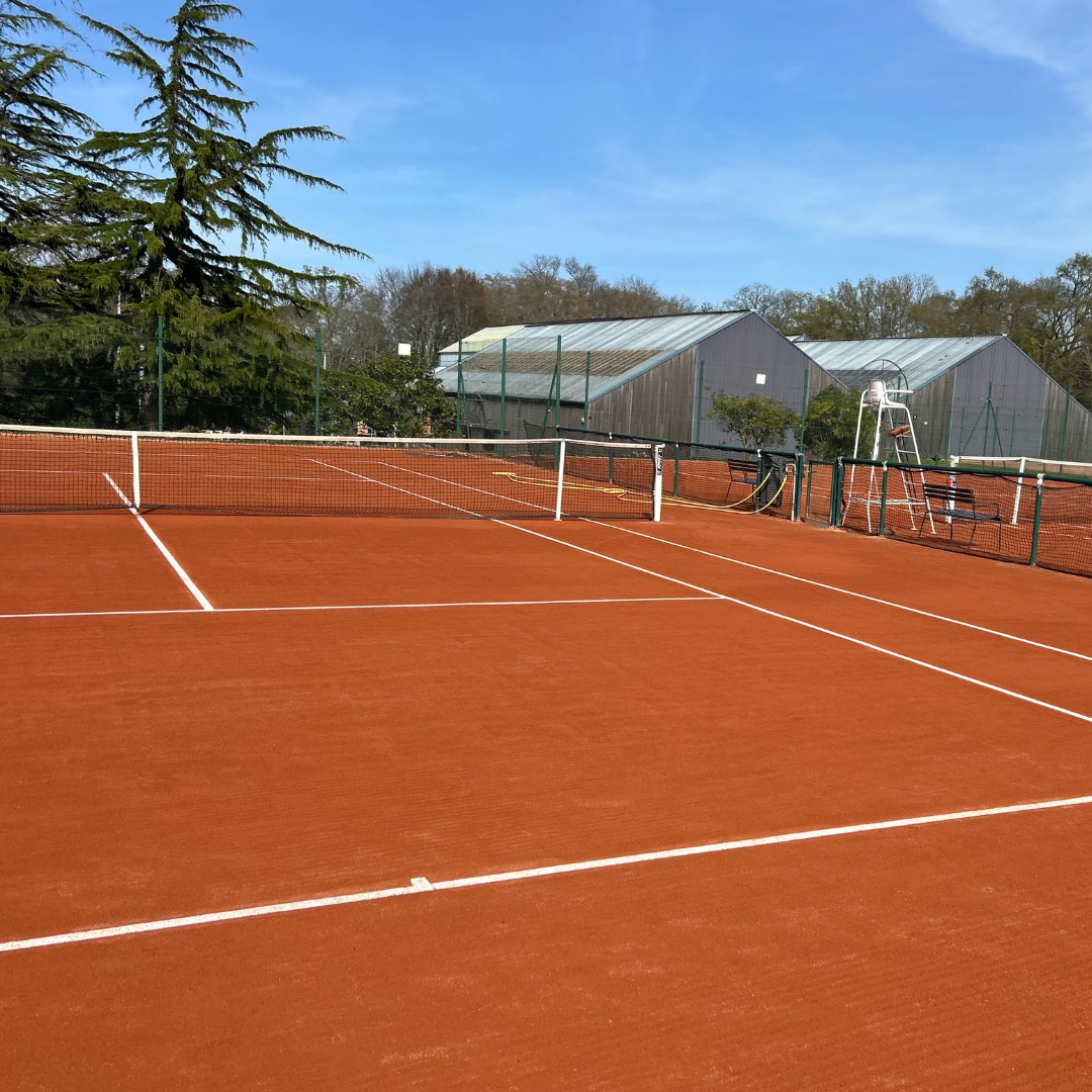 Court de tennis en terre battue avec filet et lignes blanches, entouré d'arbres, avec des bâtiments en arrière-plan et un ciel bleu.