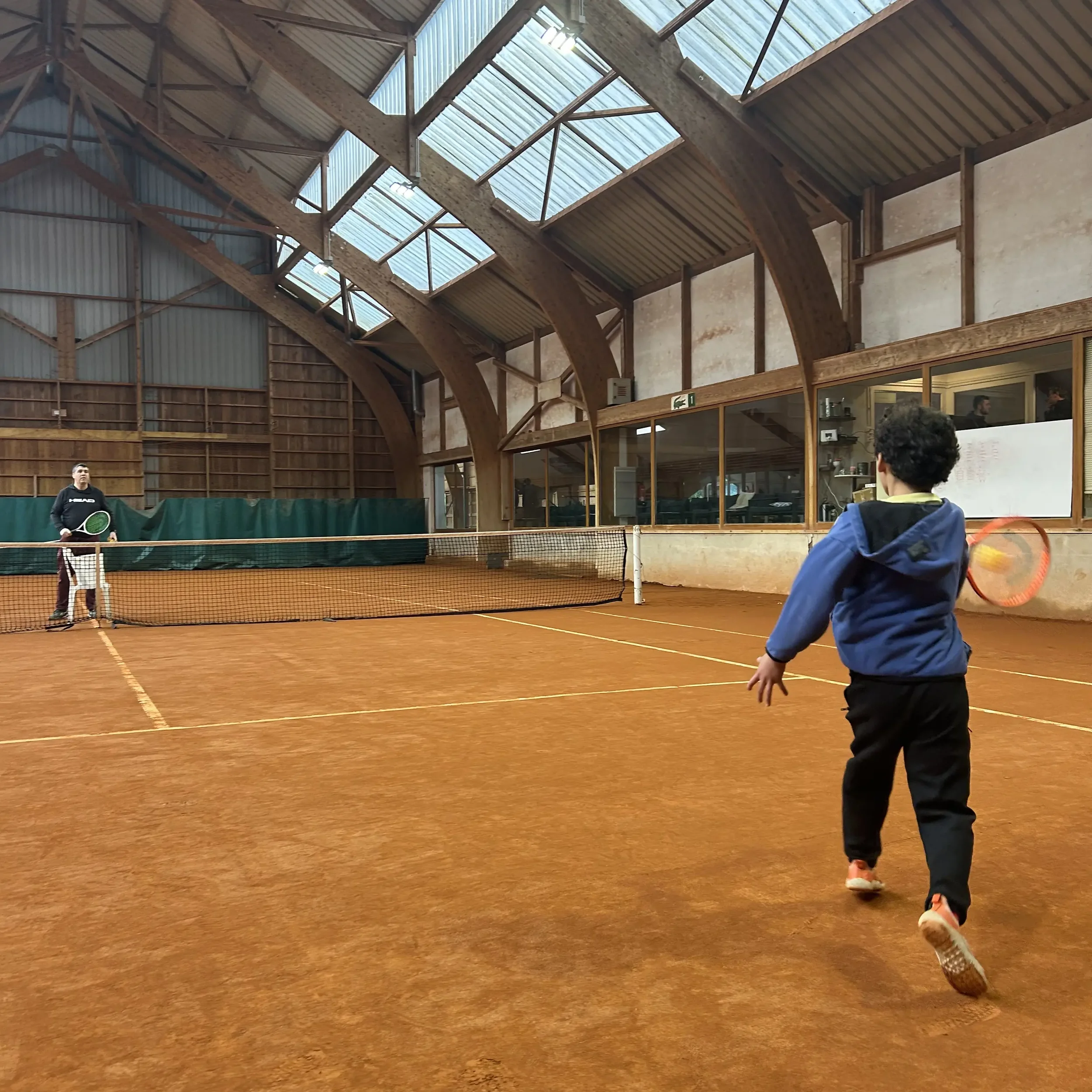Un enfant joue au tennis avec un adulte dans une salle intérieure de tennis en terre battue.