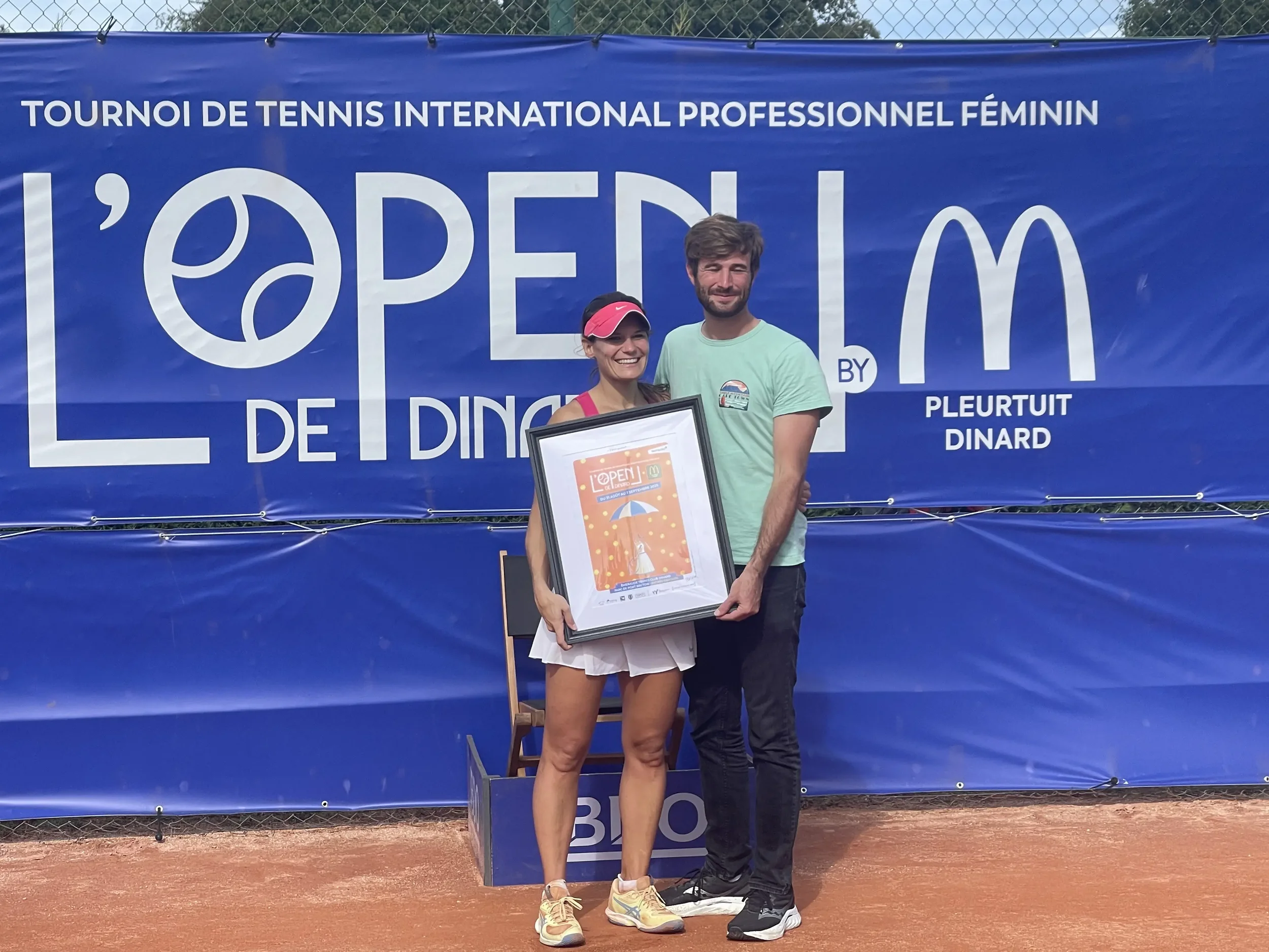 Deux personnes posent avec un cadre sur un terrain de tennis en terre battue, devant un panneau bleu avec le texte 'L'OPEN DE DINARD', une femme en tenue de sport avec visière et un homme en t-shirt vert.