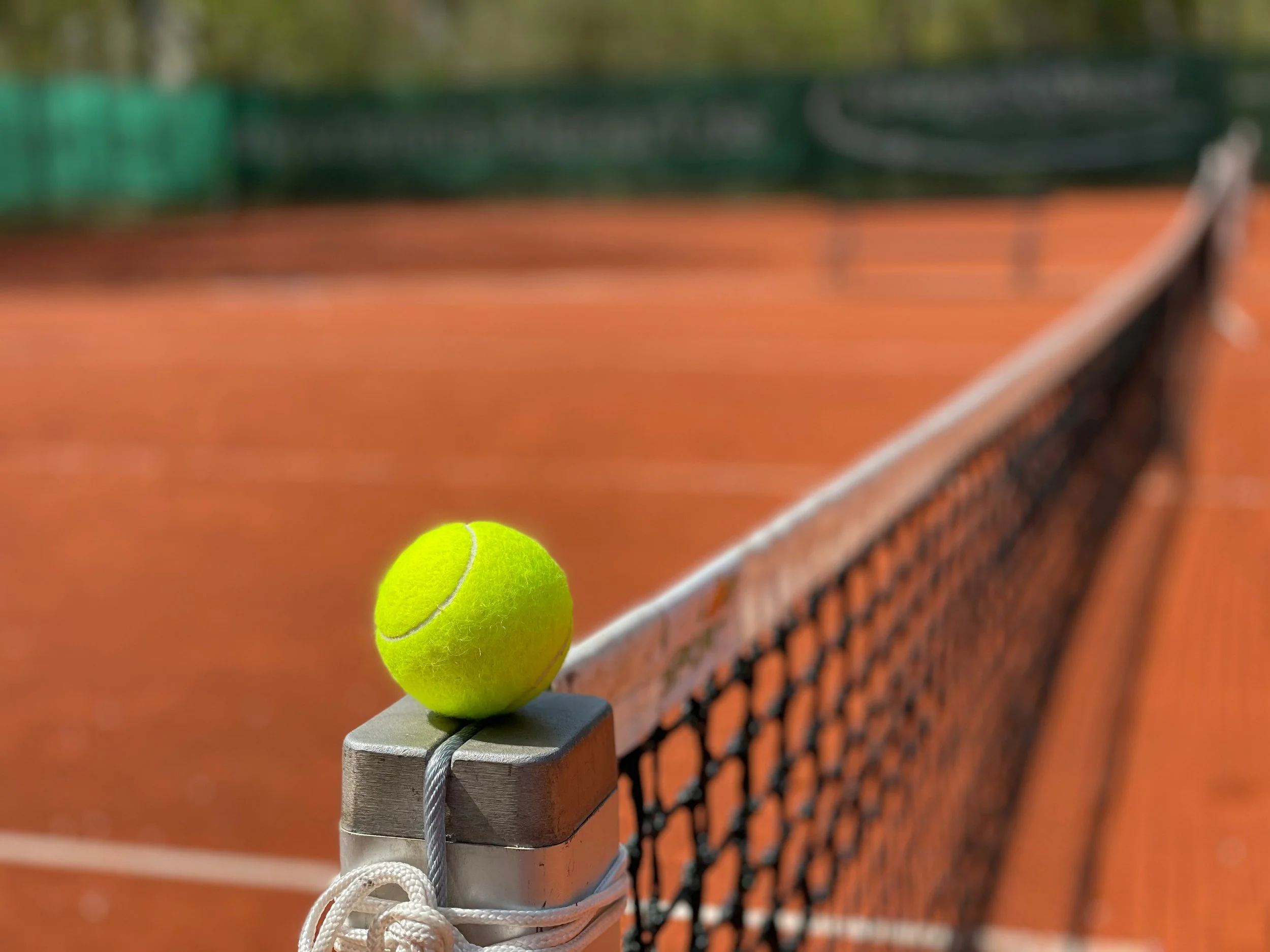 Balle de tennis jaune posée sur la tête d'un poteau de filet de tennis sur un terrain en terre battue.