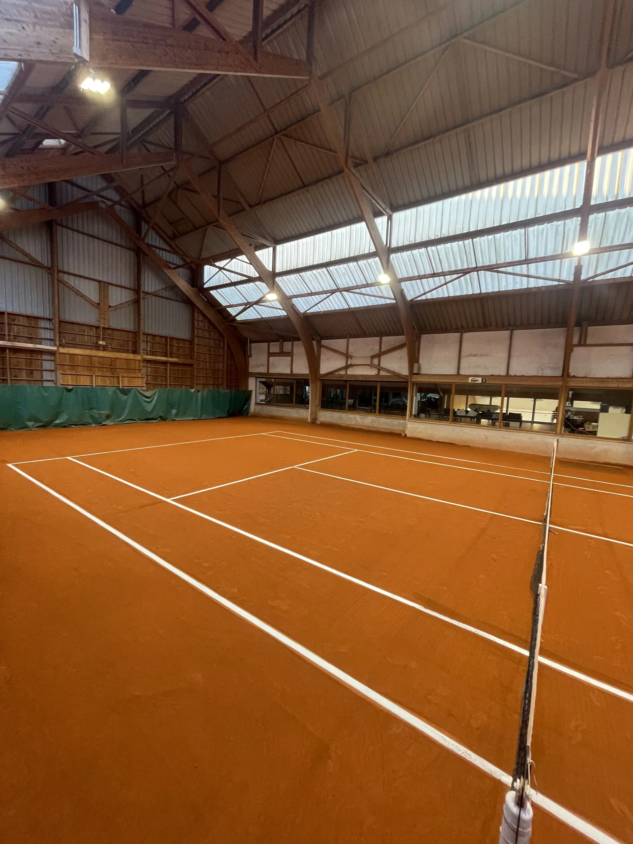 Un court de tennis en terre battue intérieur, avec un plafond en bois et des lumières suspendues.