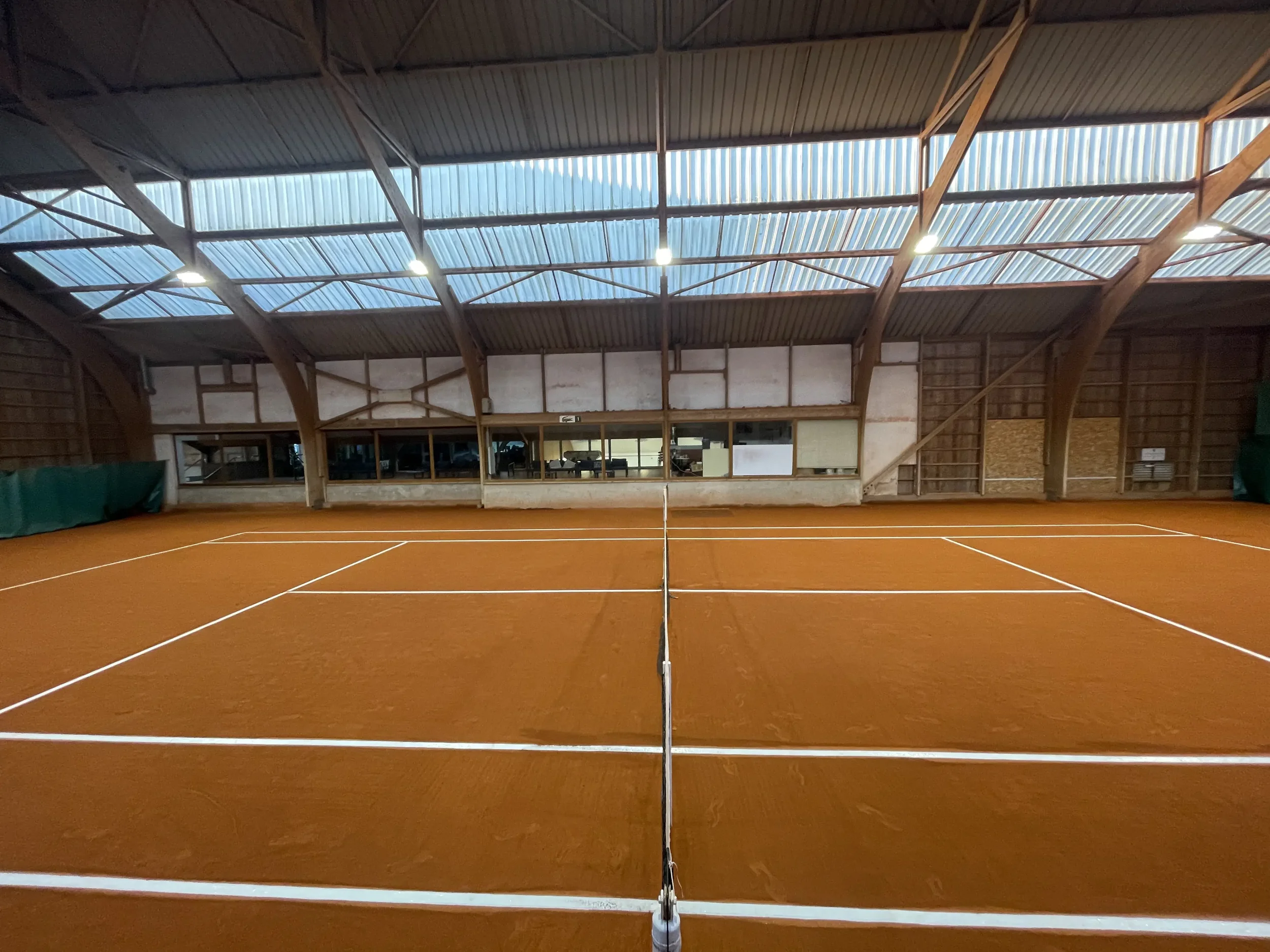 Court de tennis intérieur en terre battue, avec un plafond en bois et un mur en arrière-plan.