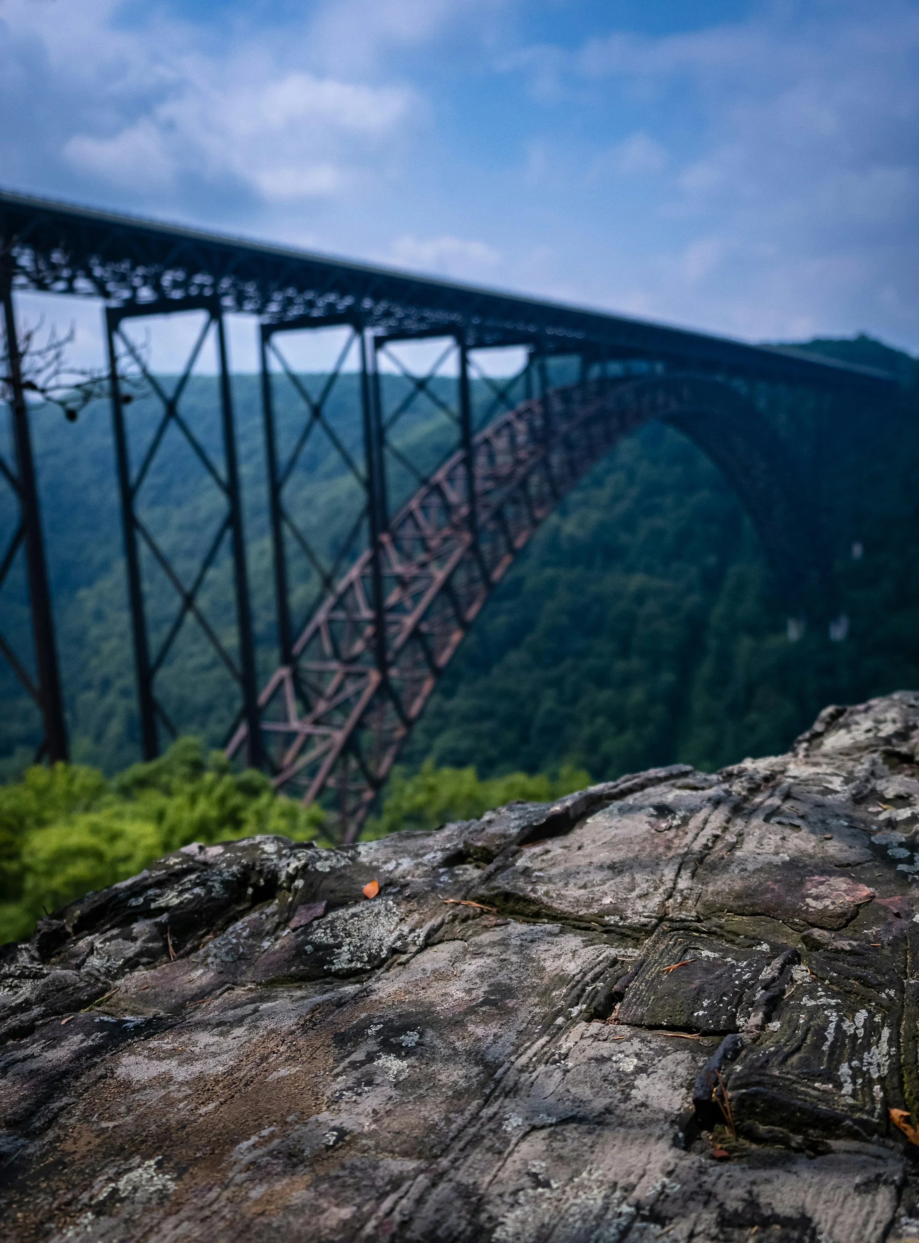 Close-up of a rock in the foreground with a blurred view of a large arch bridge and green mountain landscape in the background under a partly cloudy sky.